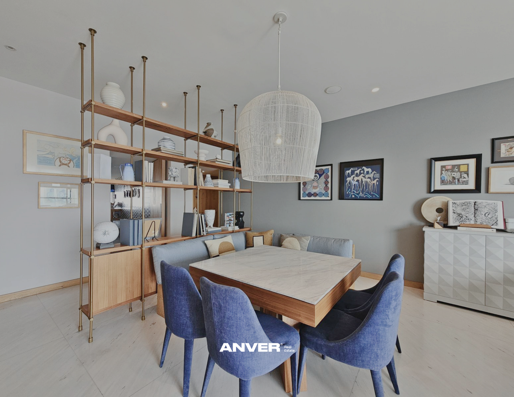 Sala de comedor moderna con mesa de mármol y sillas azul marino, estantería de madera y metal con decoraciones y libros, cuadros en la pared y lámpara de techo de mimbre.
