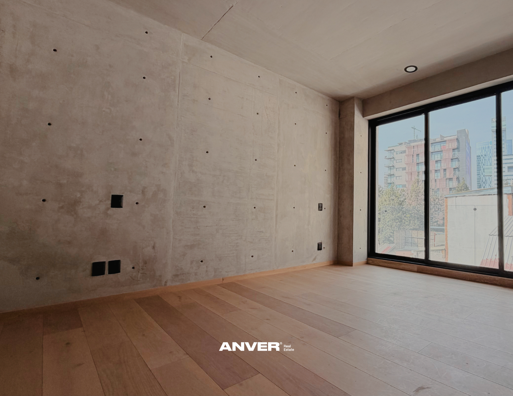 Habitación vacía en un edificio moderno con piso de madera, pared de concreto con orificios y vista a edificios urbanos a través de un gran ventanal.