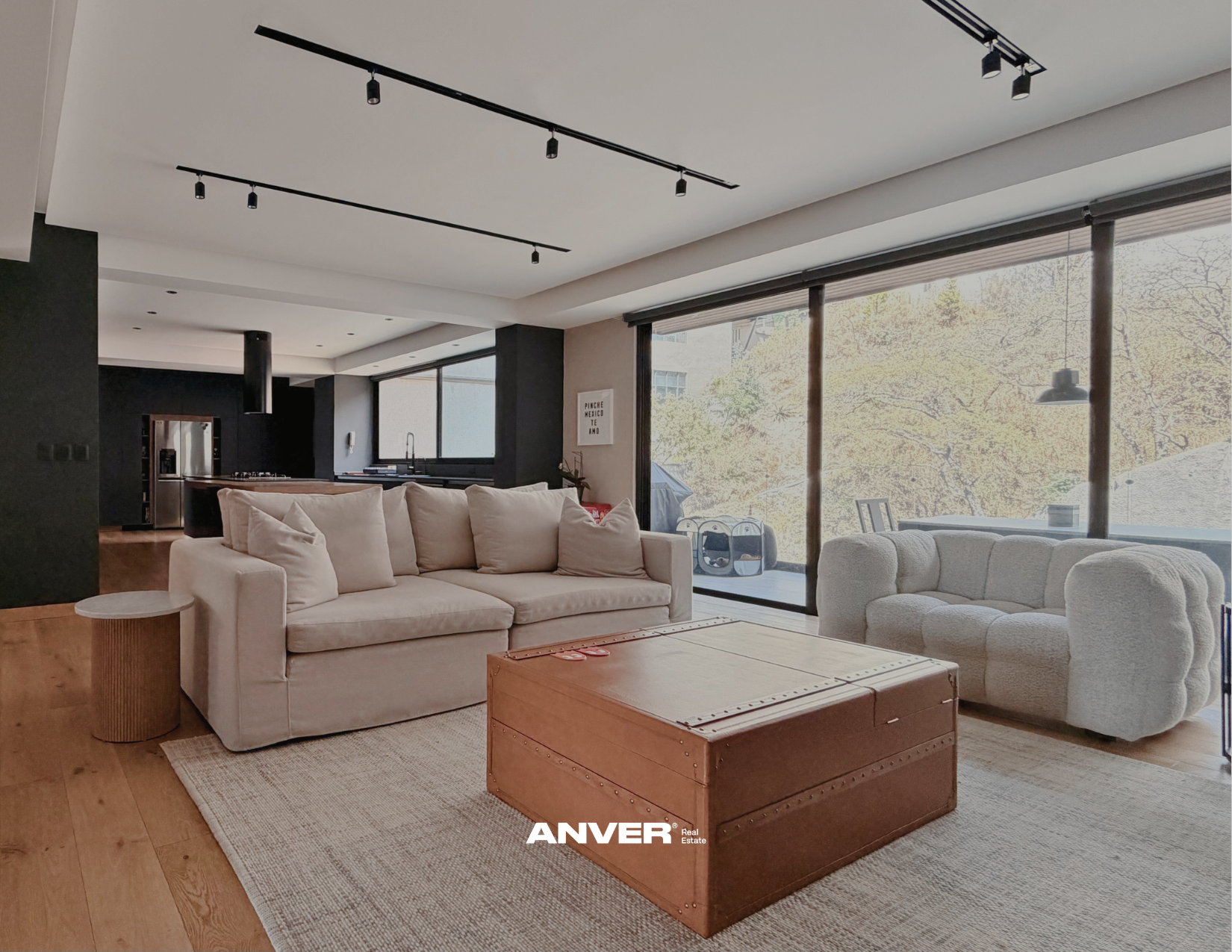 Sala de estar moderna con sofá beige, sillón blanco, mesa de madera y vista al exterior con ventanas grandes y árboles
