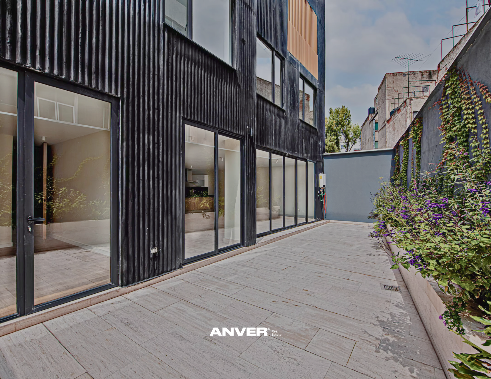 Vista de una fachada moderna de un edificio con ventanas grandes de marco negro y una entrada de puerta de vidrio. Jardinera con plantas verdes y flores moradas a la derecha, y piso de piedra clara en el patio. Cielo parcialmente nublado con árboles al fondo.