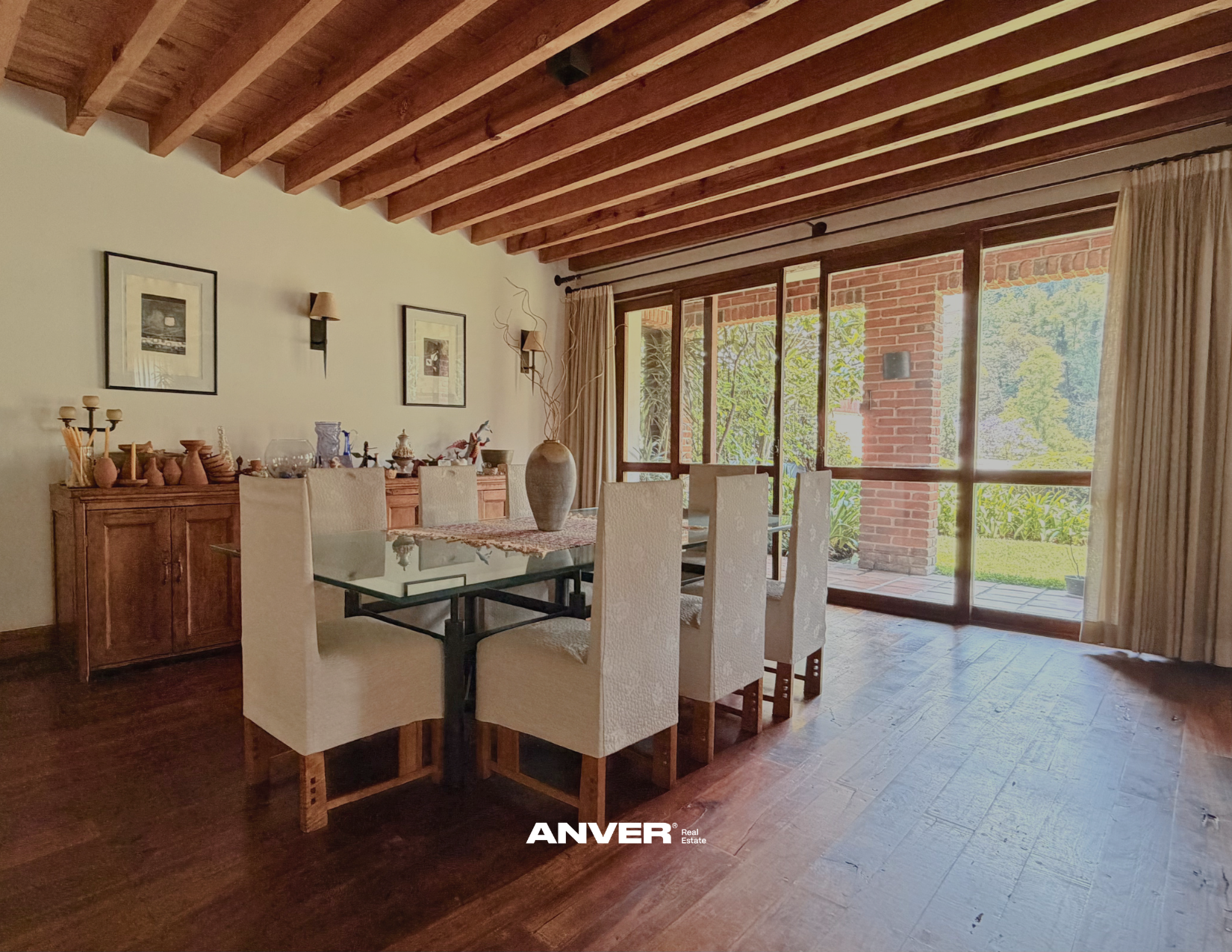 Comedor con mesa de cristal y sillas blancas, vista a balcón con ventanas grandes y puertas de vidrio, decoración con cuadros, armario de madera y jarrón con ramas secas, piso de madera y techo de madera con vigas.