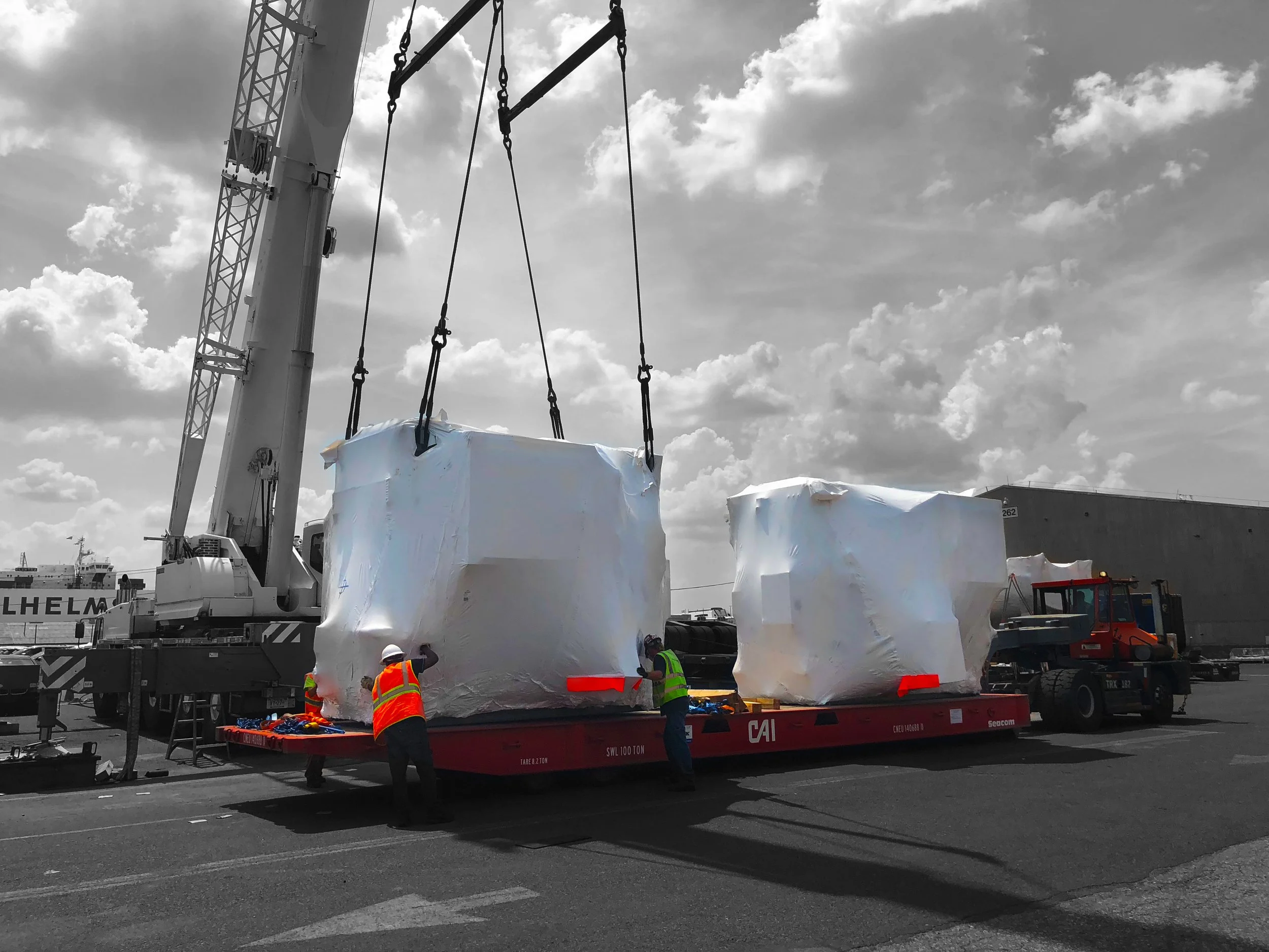Large industrial equipment or possibly a spacecraft component covered in white protective material being unloaded from a flatbed truck with workers in safety vests assisting, against a cloudy sky. We handle all kinds of high and heavy equipment
