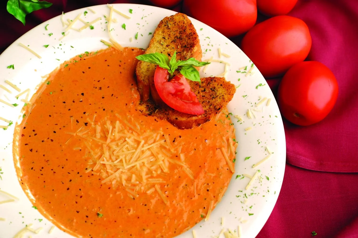 A plate of creamy tomato soup garnished with shredded cheese, black pepper, and fresh herbs. Two slices of toasted bread with a basil leaf and a slice of tomato on top are on the plate. Whole tomatoes are placed on a red cloth beside the plate.