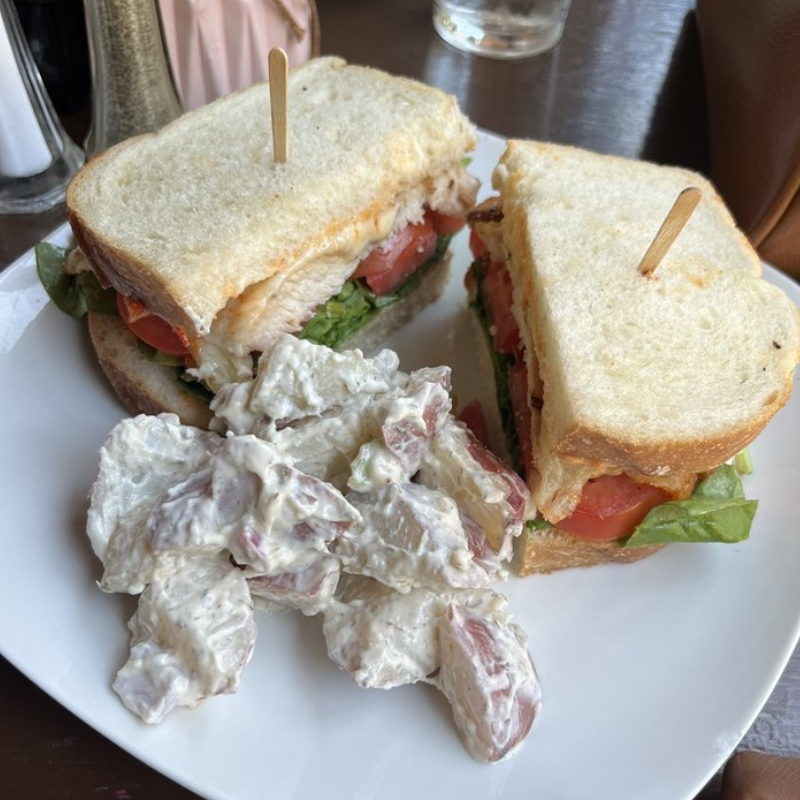 Club sandwich with layers of turkey, bacon, lettuce, tomato, and mayonnaise on toasted white bread, served with a side of potato salad.