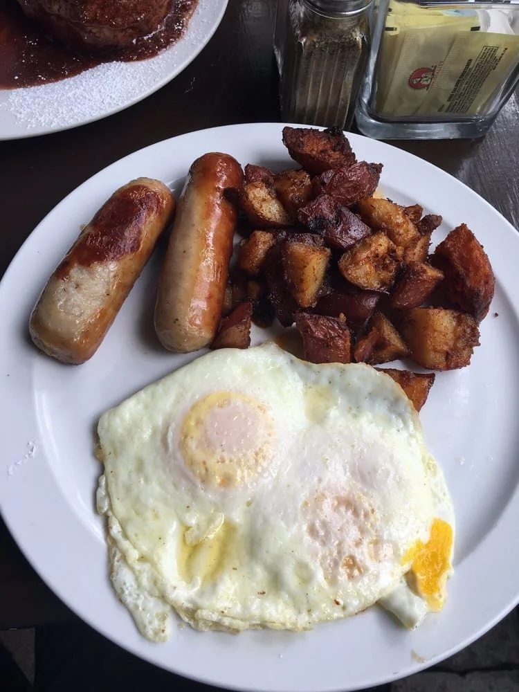 Breakfast plate with two fried eggs, two sausage links, and diced crispy potatoes, served on a white plate.