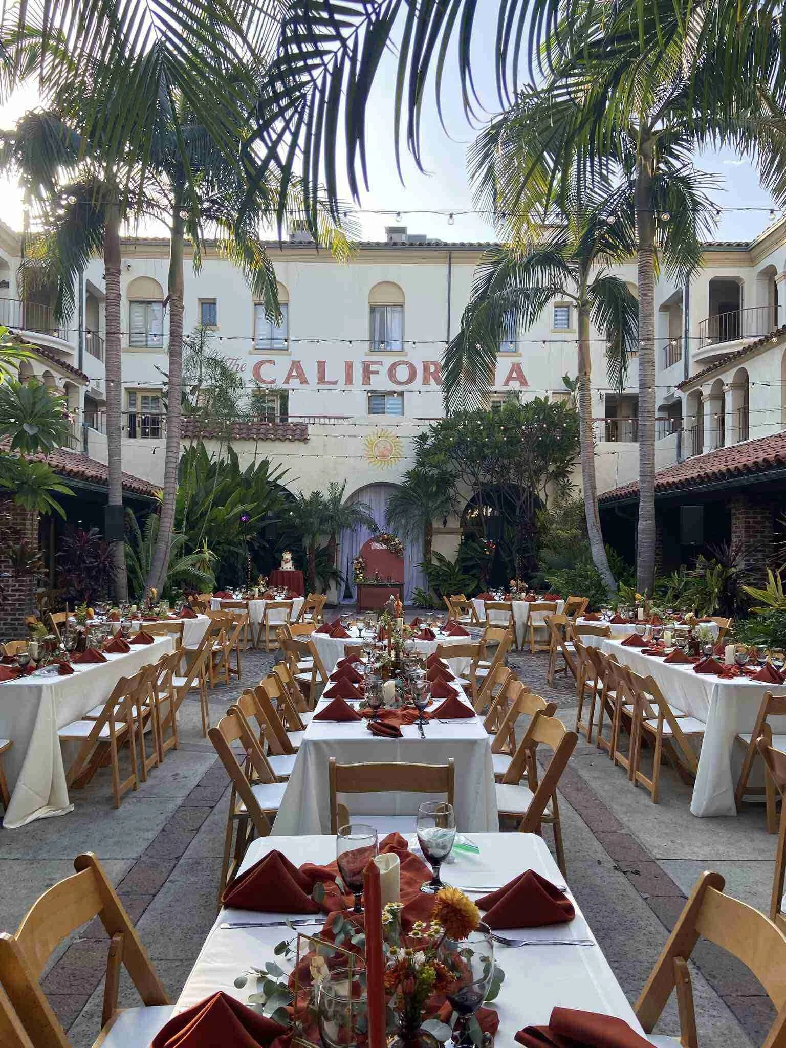 An outdoor courtyard decorated for a celebration with tables set for a meal, each with white tablecloths, folded red napkins, flowers, and candles. Tall palm trees line the space, and a building in the background has a sign reading 'The California' w
