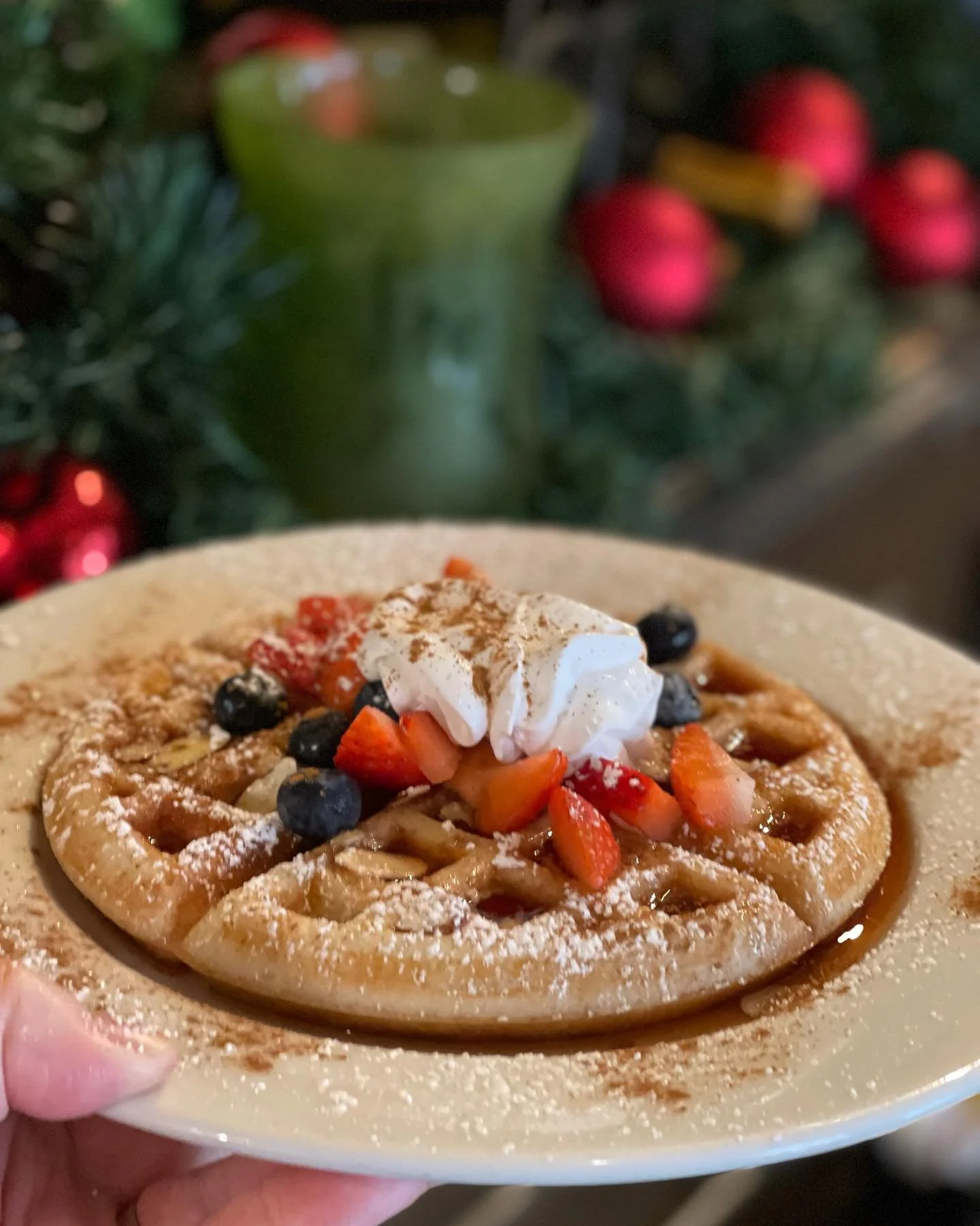 A waffle topped with whipped cream, strawberries, blueberries, and a dusting of powdered sugar, served on a beige plate.