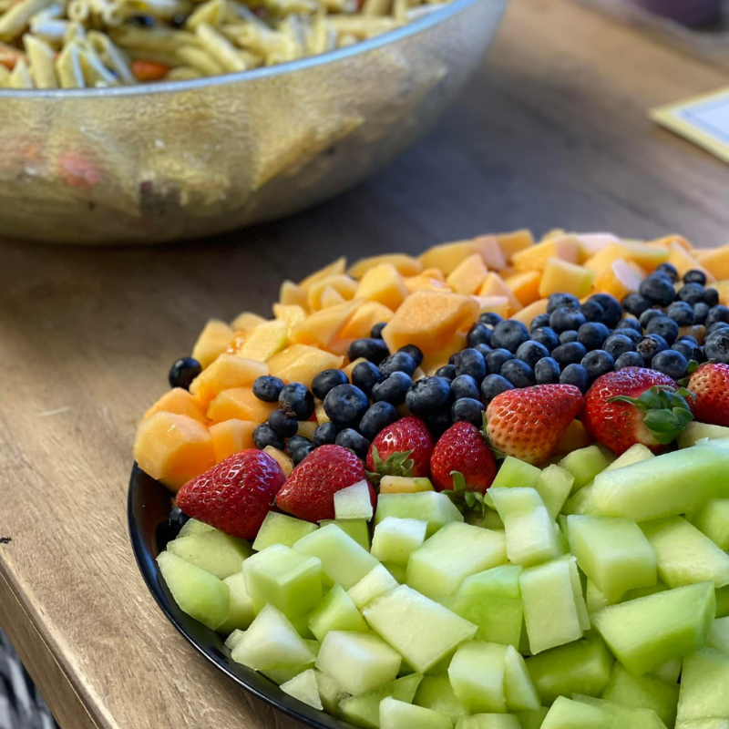 Plate of fresh cut cantaloupe, strawberries, blueberries, and honeydew melon, with a large bowl of pasta salad nearby on a wooden table.