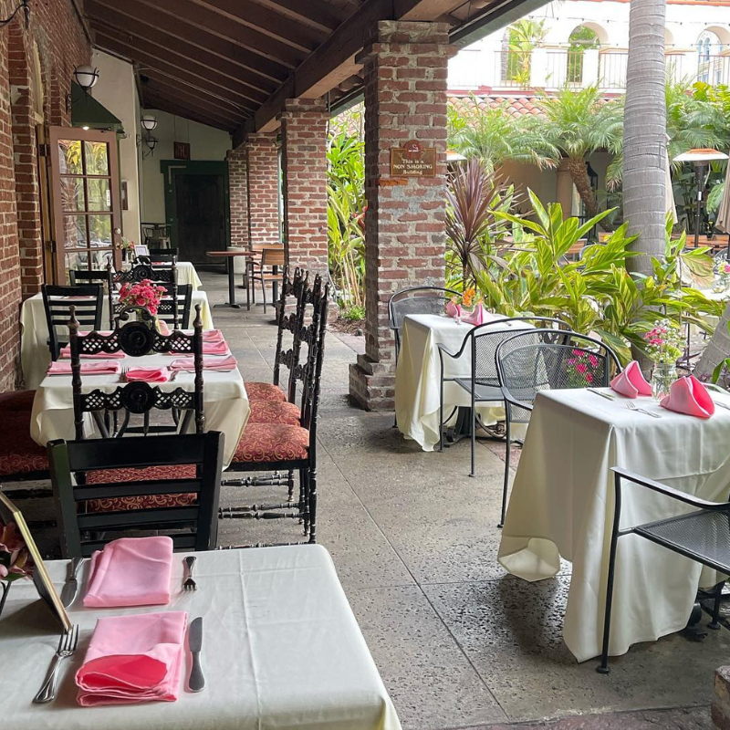 Outdoor restaurant patio with brick columns, white tablecloths, pink napkins, black and patterned chairs, and lush green plants.