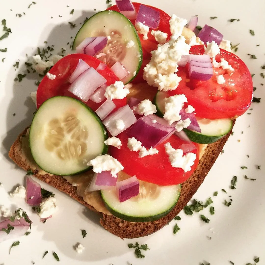 Open-faced sandwich with sliced cucumbers, tomatoes, red onions, crumbled feta cheese, on whole grain bread, garnished with chopped herbs.