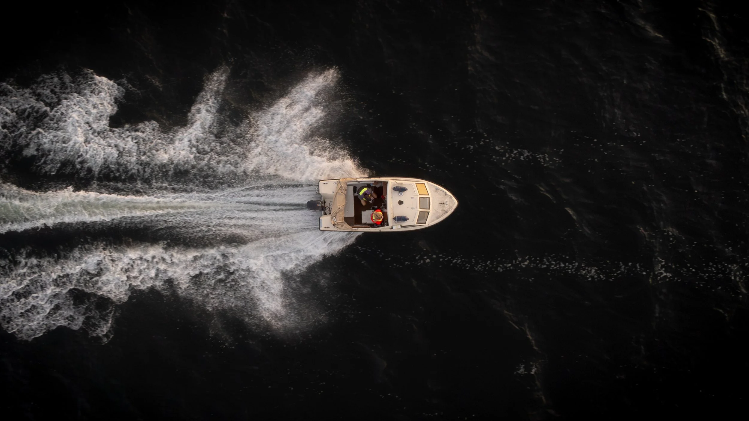 Aerial view of a small boat with two people on dark water, creating a wake as it moves.