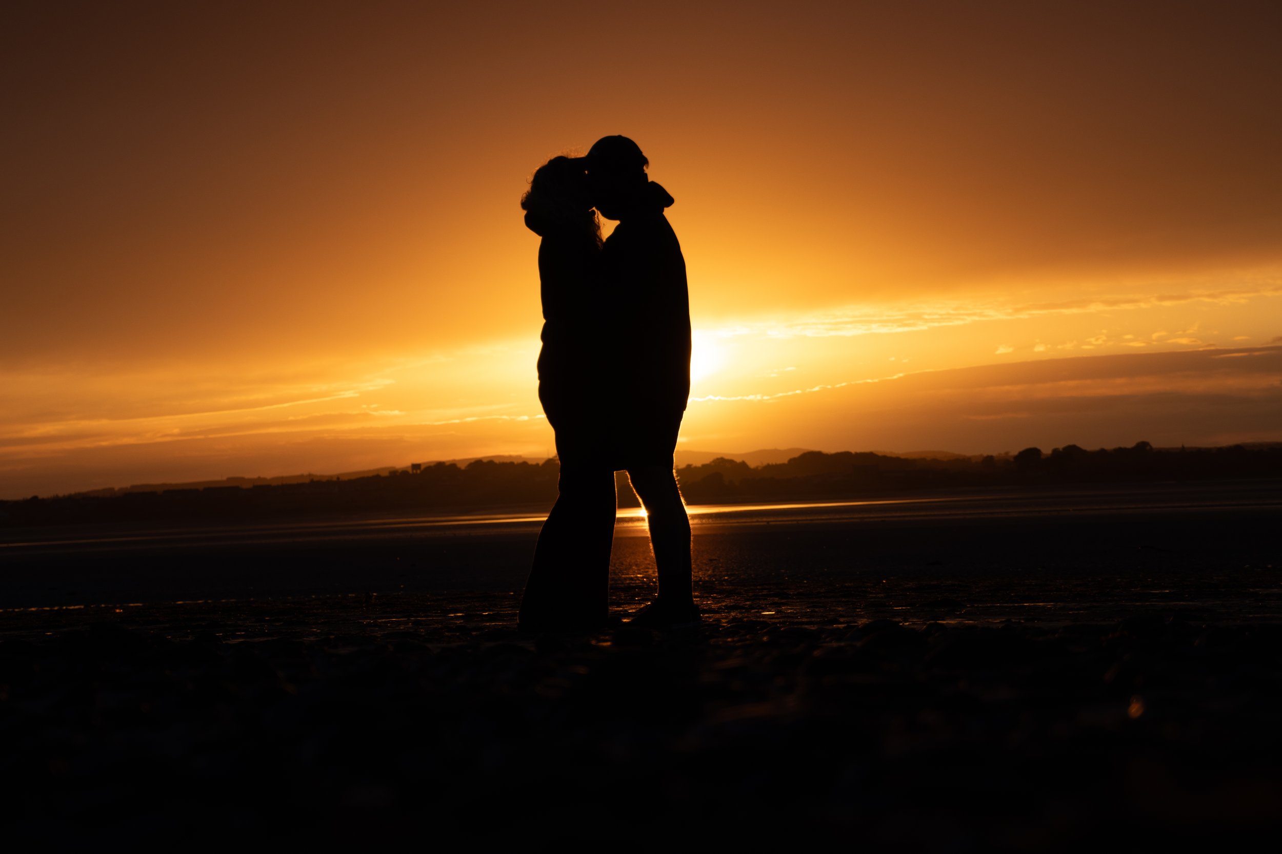 Silhouetted couple kissing at sunset on the beach, with the ocean in the background and golden sky.