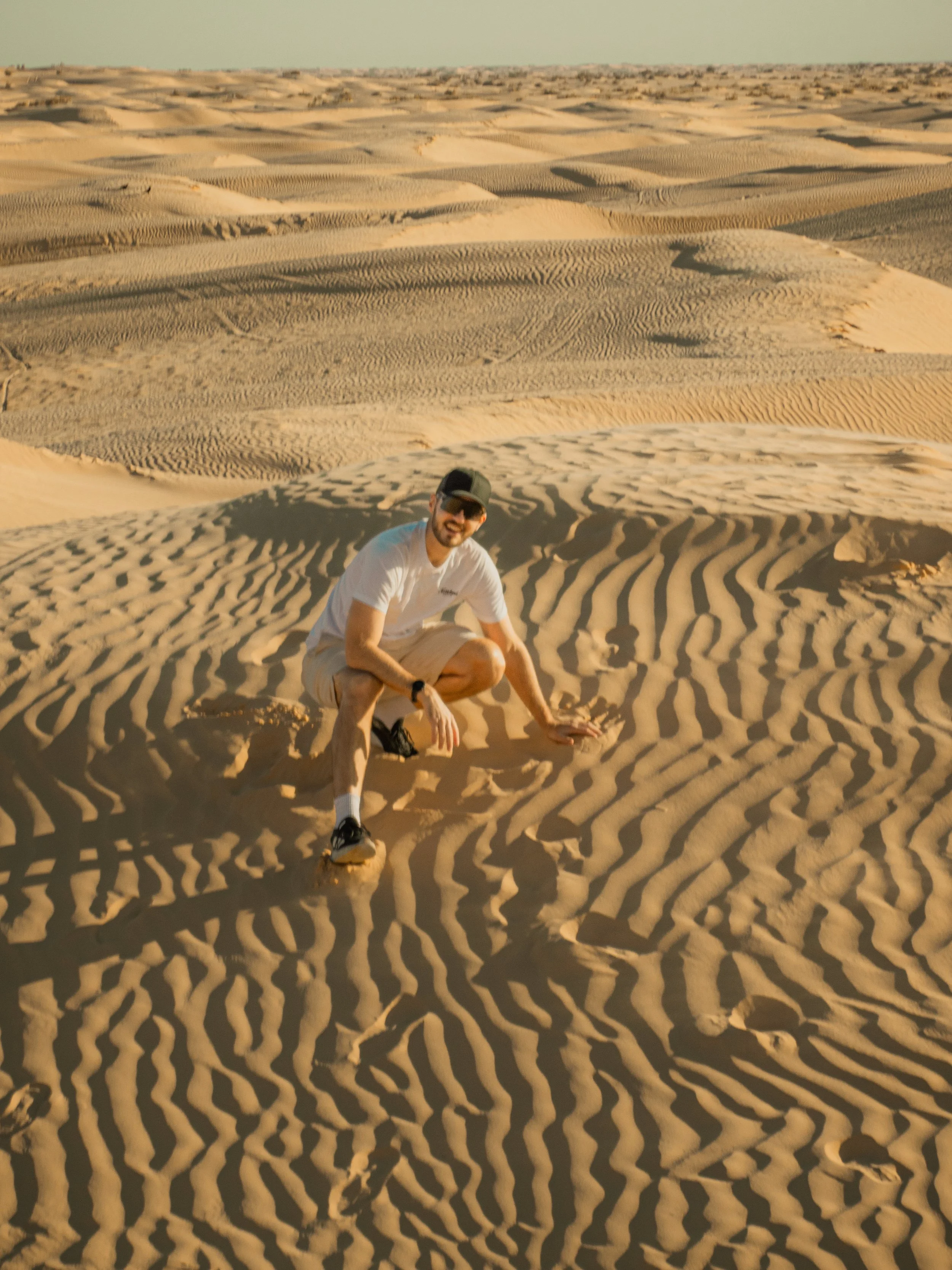 A man in a white T-shirt, shorts, black cap, and sunglasses is crouching on sand dunes in a desert, with a vast expanse of rippled sand dunes extending to the horizon under a clear sky.