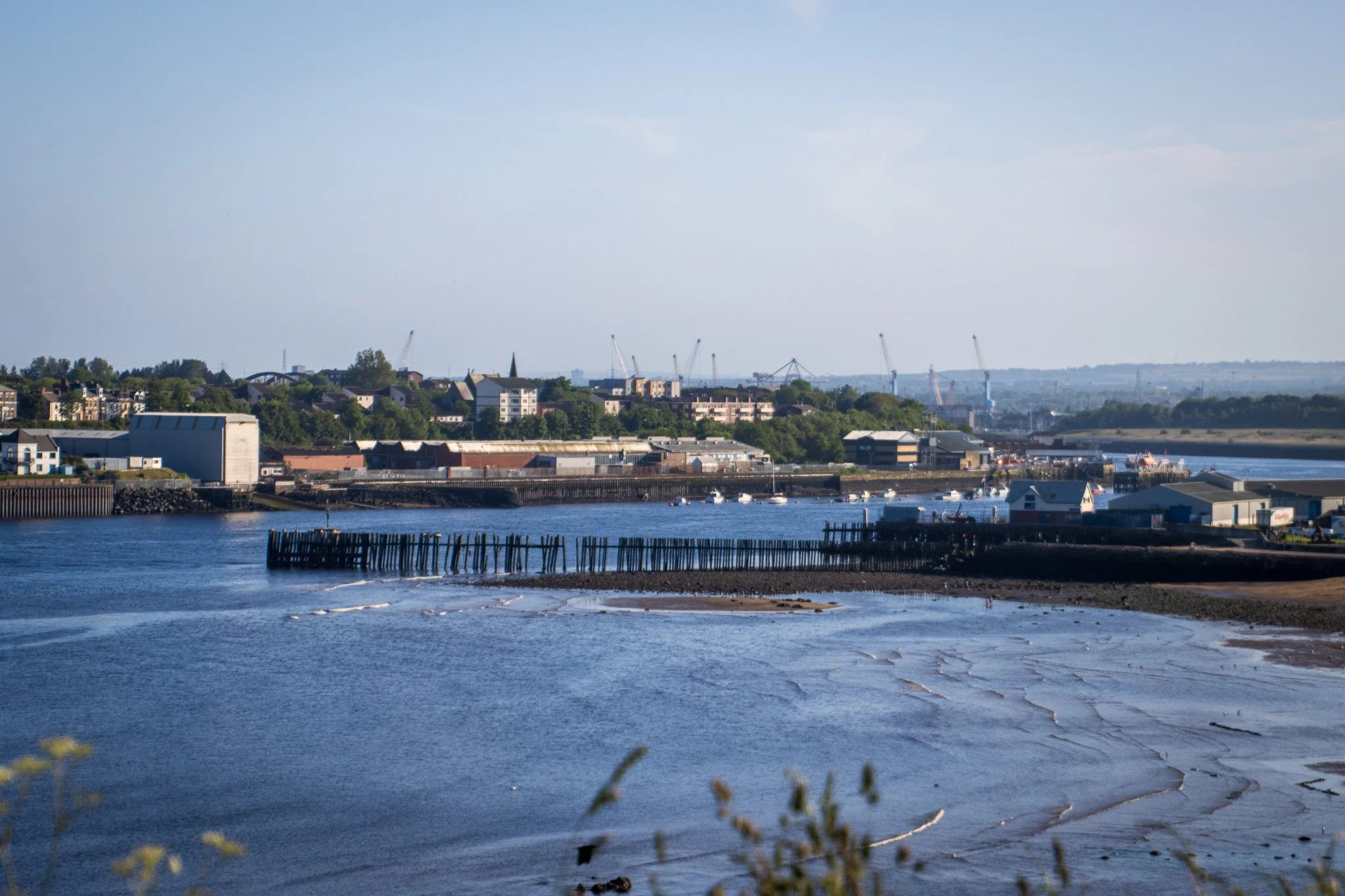 A scenic view of a harbor with boats docked, industrial buildings, and a hillside with houses and cranes under a clear sky.