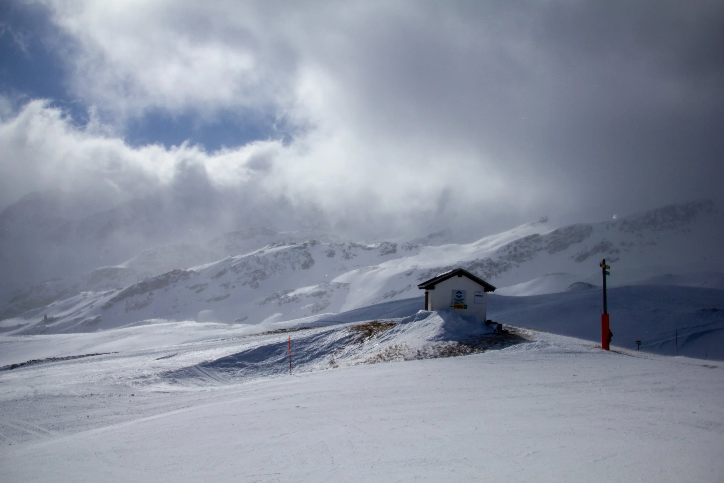 Snow-covered landscape with a mountain in the background, partly obscured by clouds and fog. A small building with signs, and a red pole in the foreground. The sky is cloudy with patches of blue showing through.