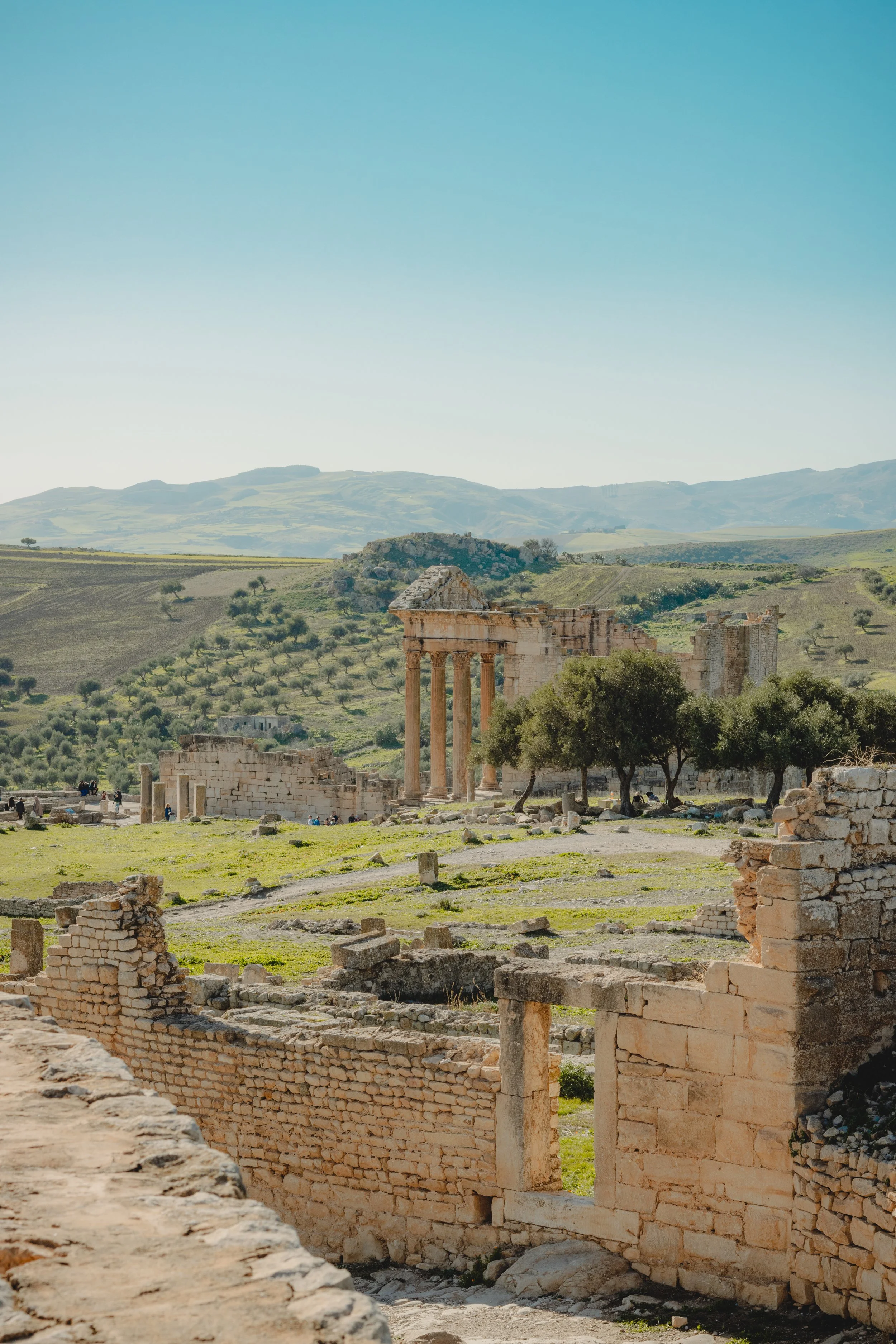Ancient ruins with upright stone columns and crumbling brick walls set in a green landscape with rolling hills in the background under a clear blue sky.