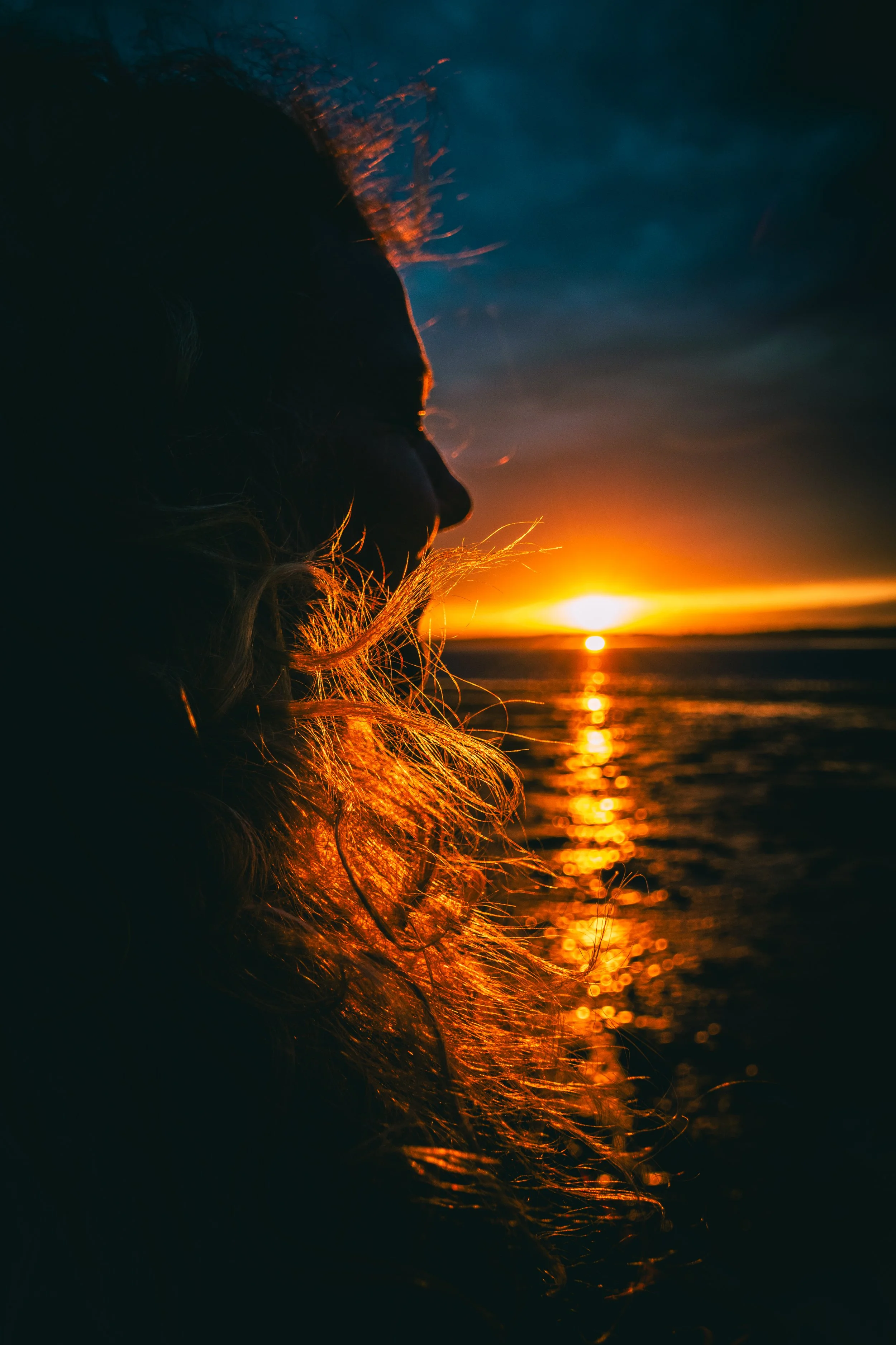 Silhouette of a person with long hair at sunset by the water, with the sun near the horizon and a dark sky overhead.