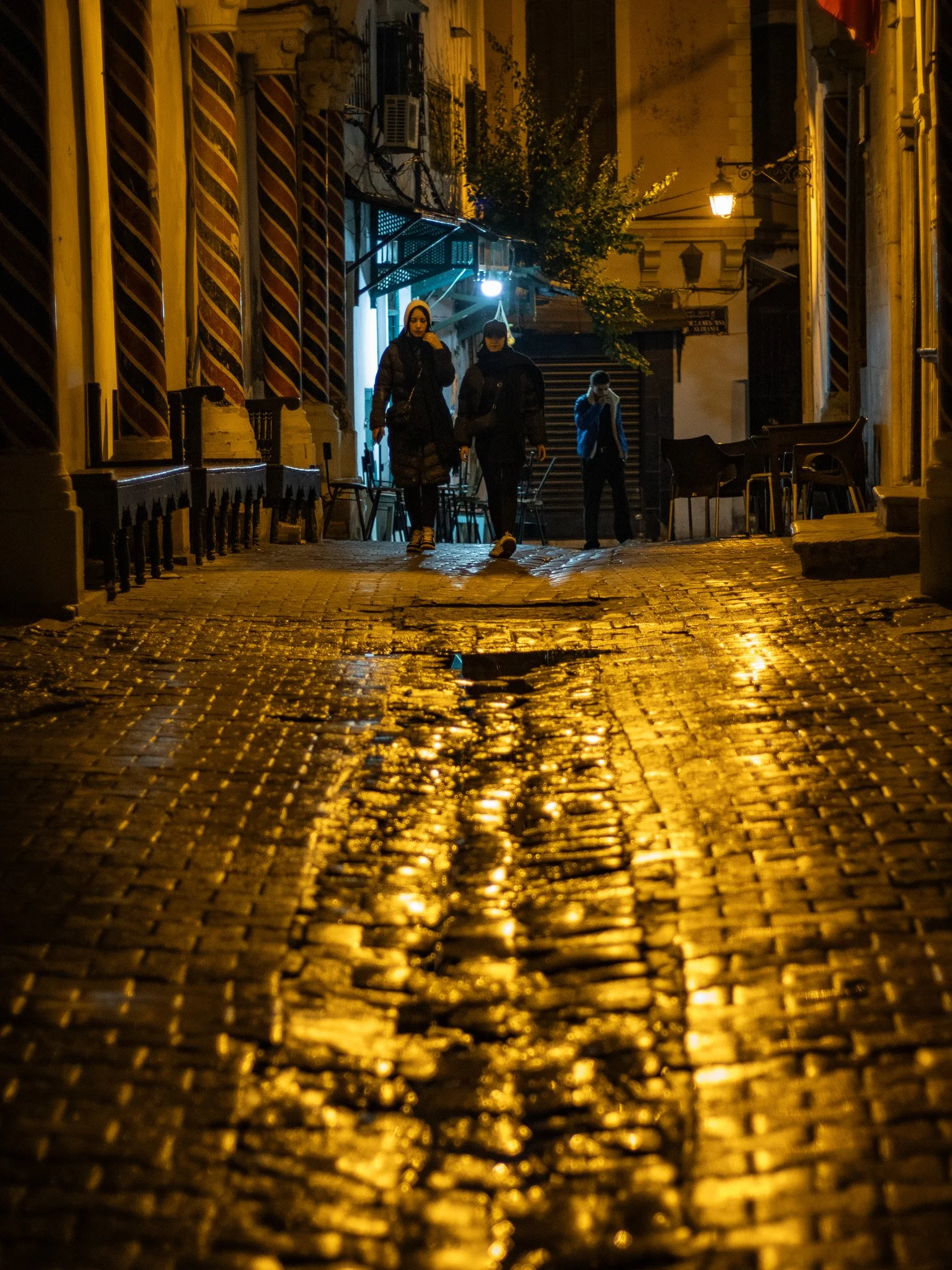 Night scene in a narrow, cobblestone street with three people walking, illuminated by warm streetlights, with chairs and tables along the sides and buildings with shutters and air conditioning units.
