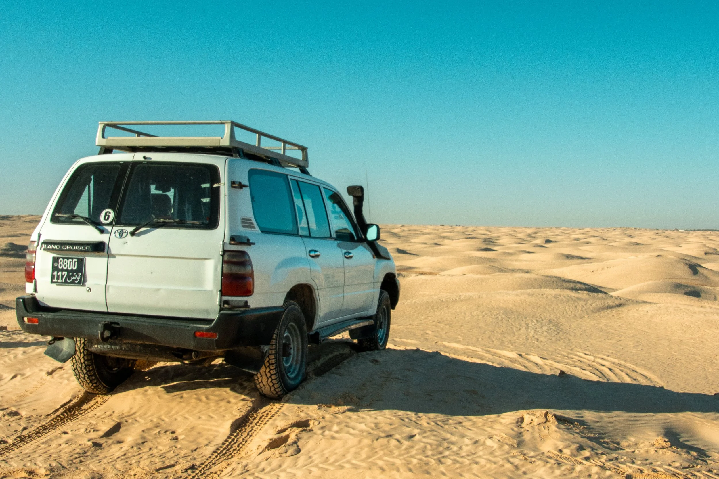 A white Toyota Land Cruiser SUV with a roof rack parked on a sandy desert terrain under a clear blue sky.