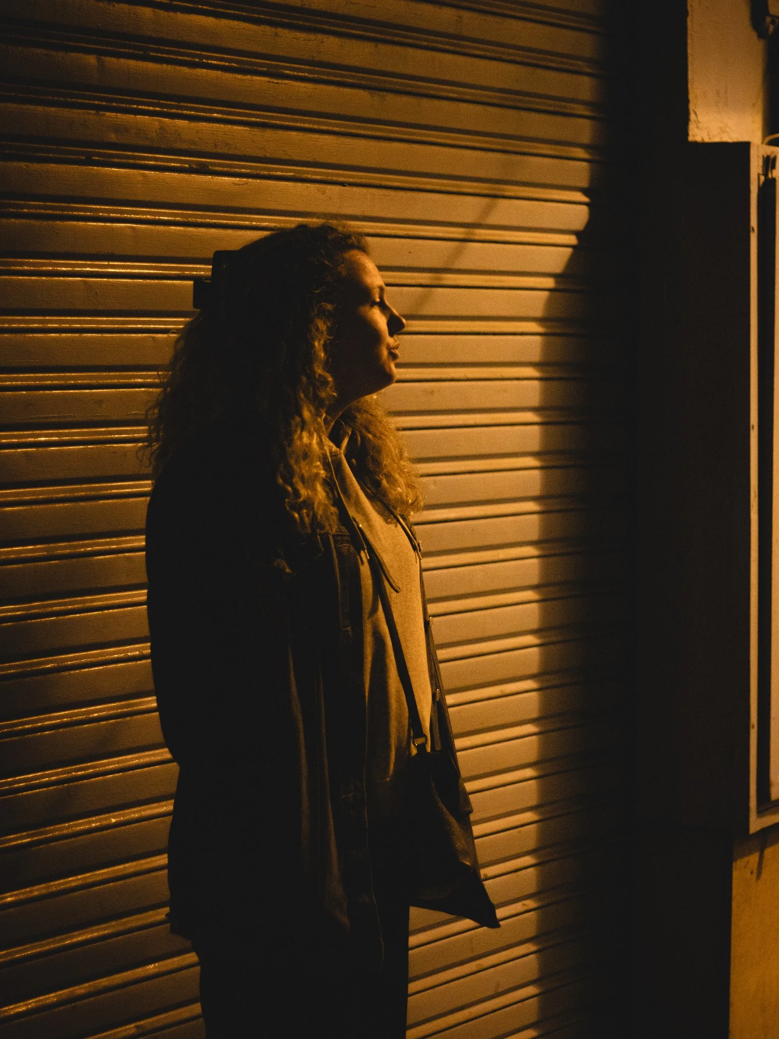 A woman with curly hair, wearing a jacket, stands with her profile visible against a closed metal shutter, looking away in a dimly lit environment.