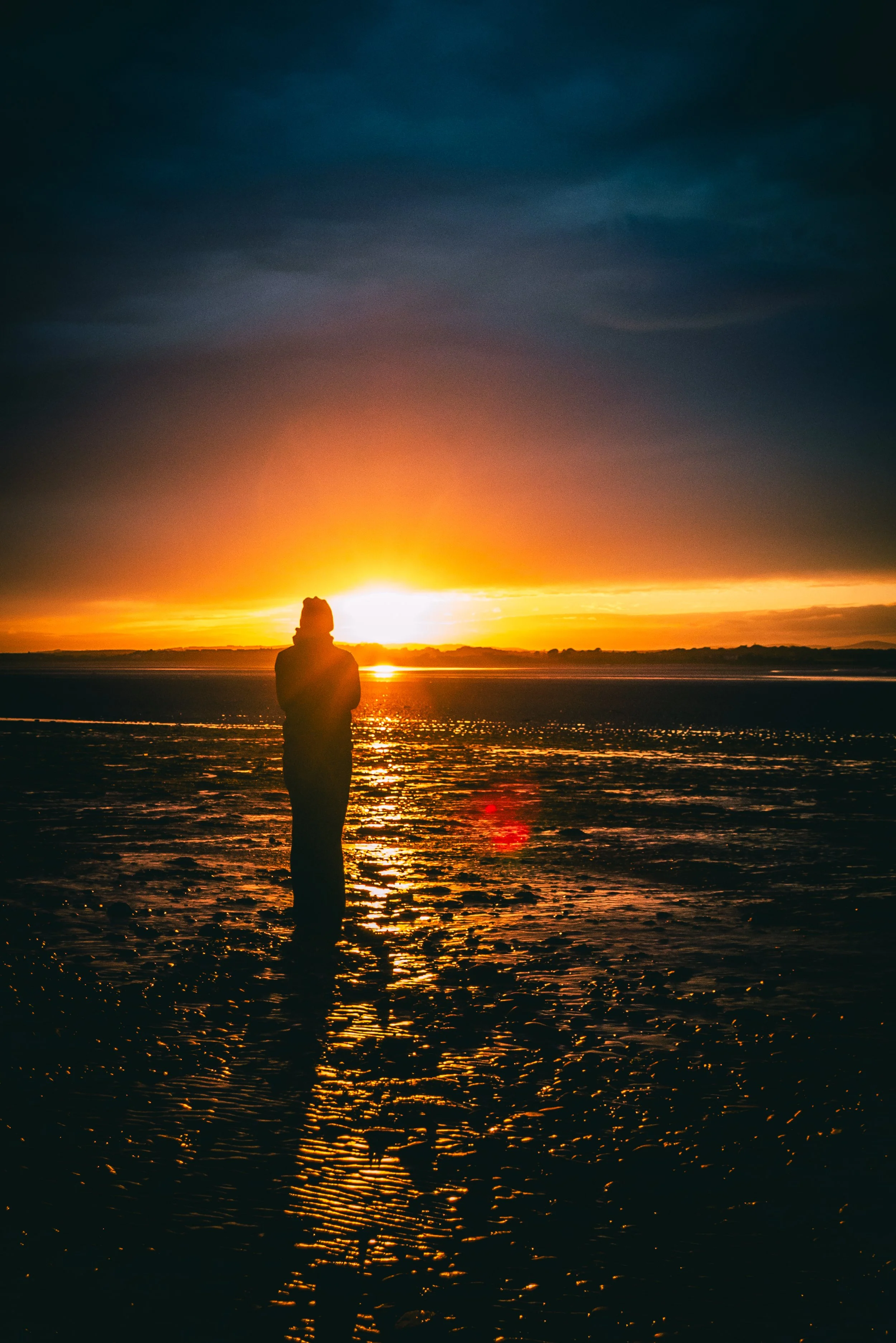 A person standing on a beach during sunset, with the sun low on the horizon and colorful clouds in the sky, reflected on the water.