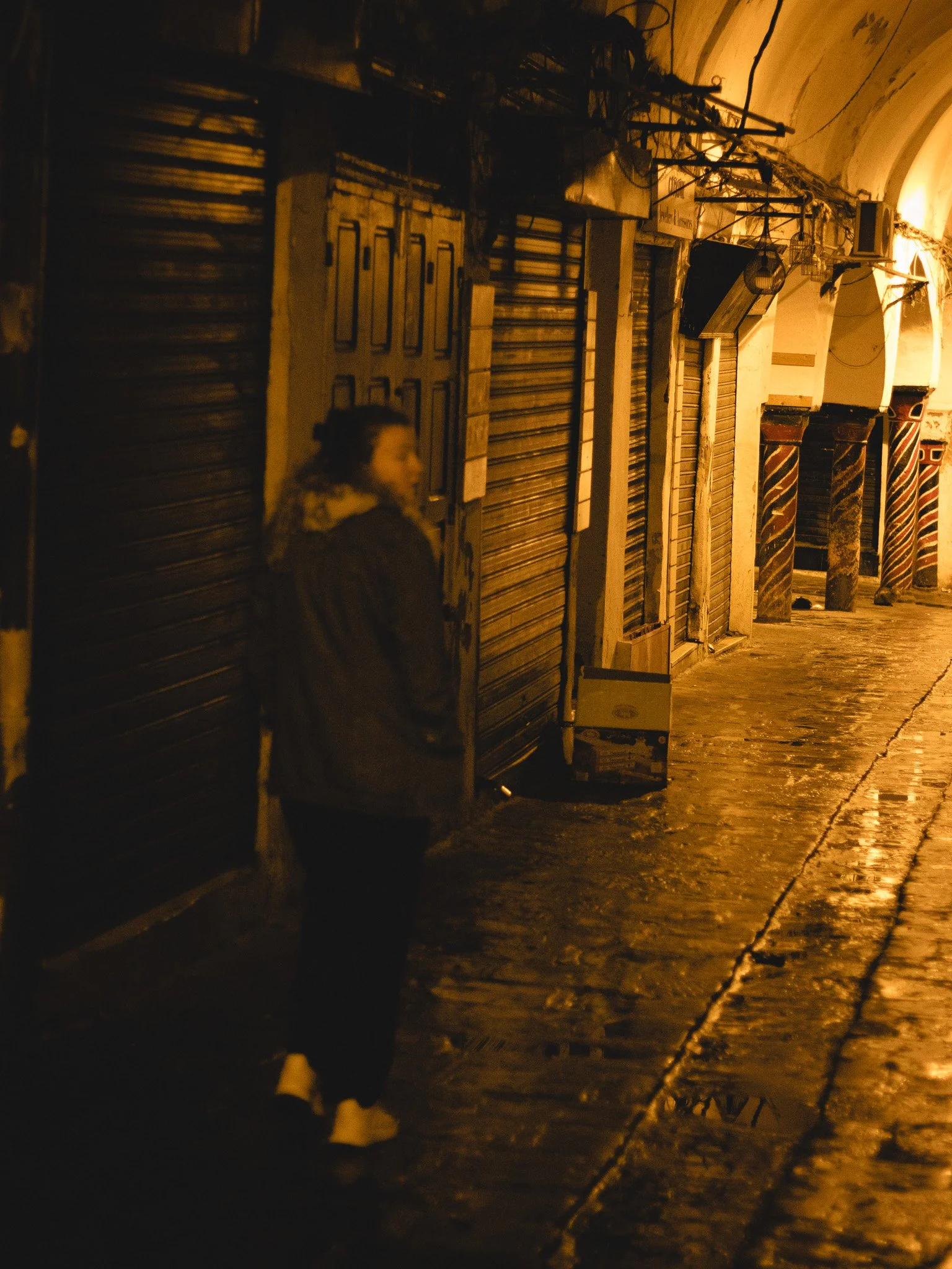 A blurry person standing on a cobblestone street at night near closed shops with metal shutters, warm lighting, and arches overhead.