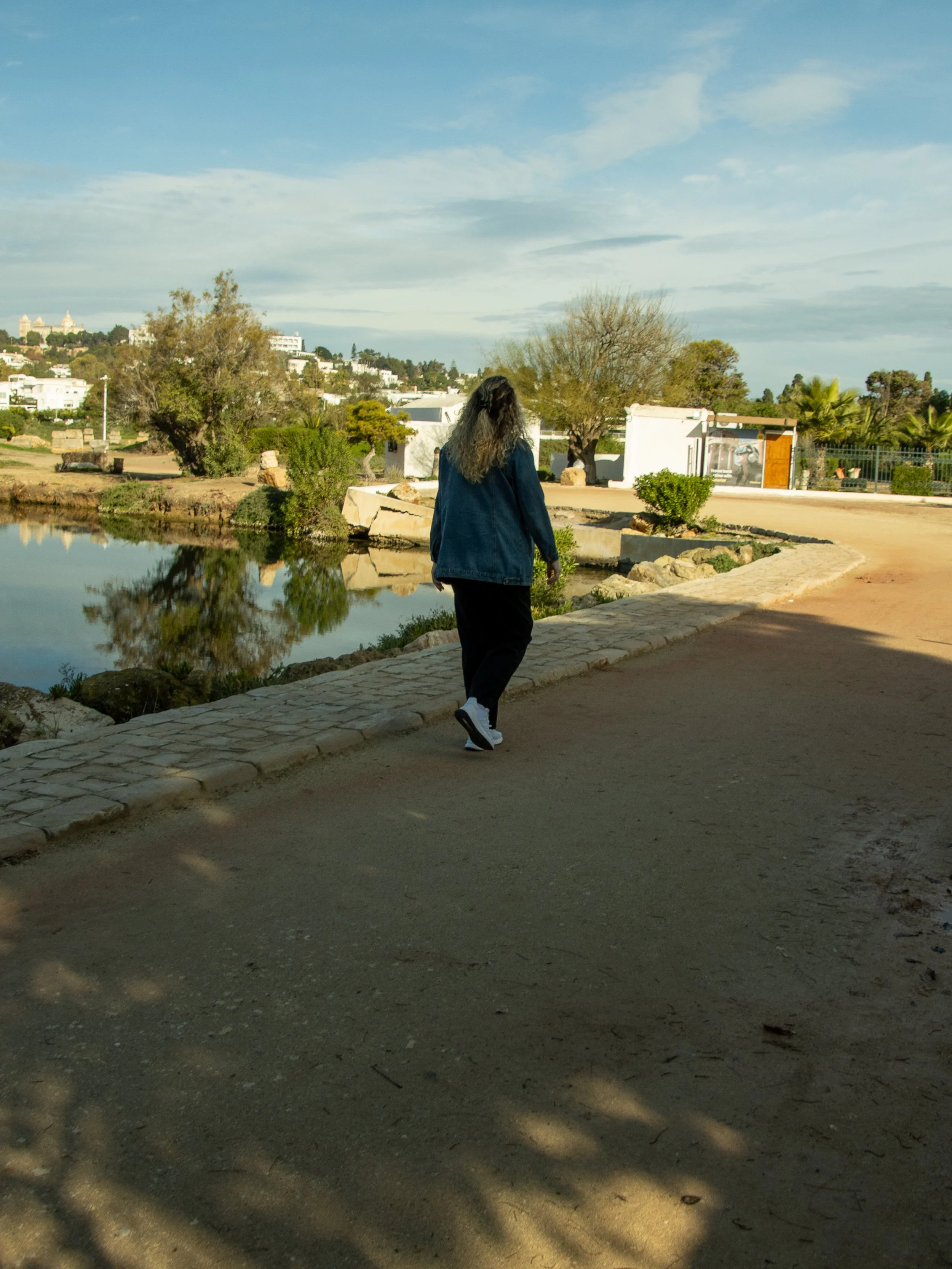 A woman with long dark hair, dressed in a blue jacket, walking along a paved path near a body of water, with trees, bushes, and buildings in the background under a partly cloudy sky.