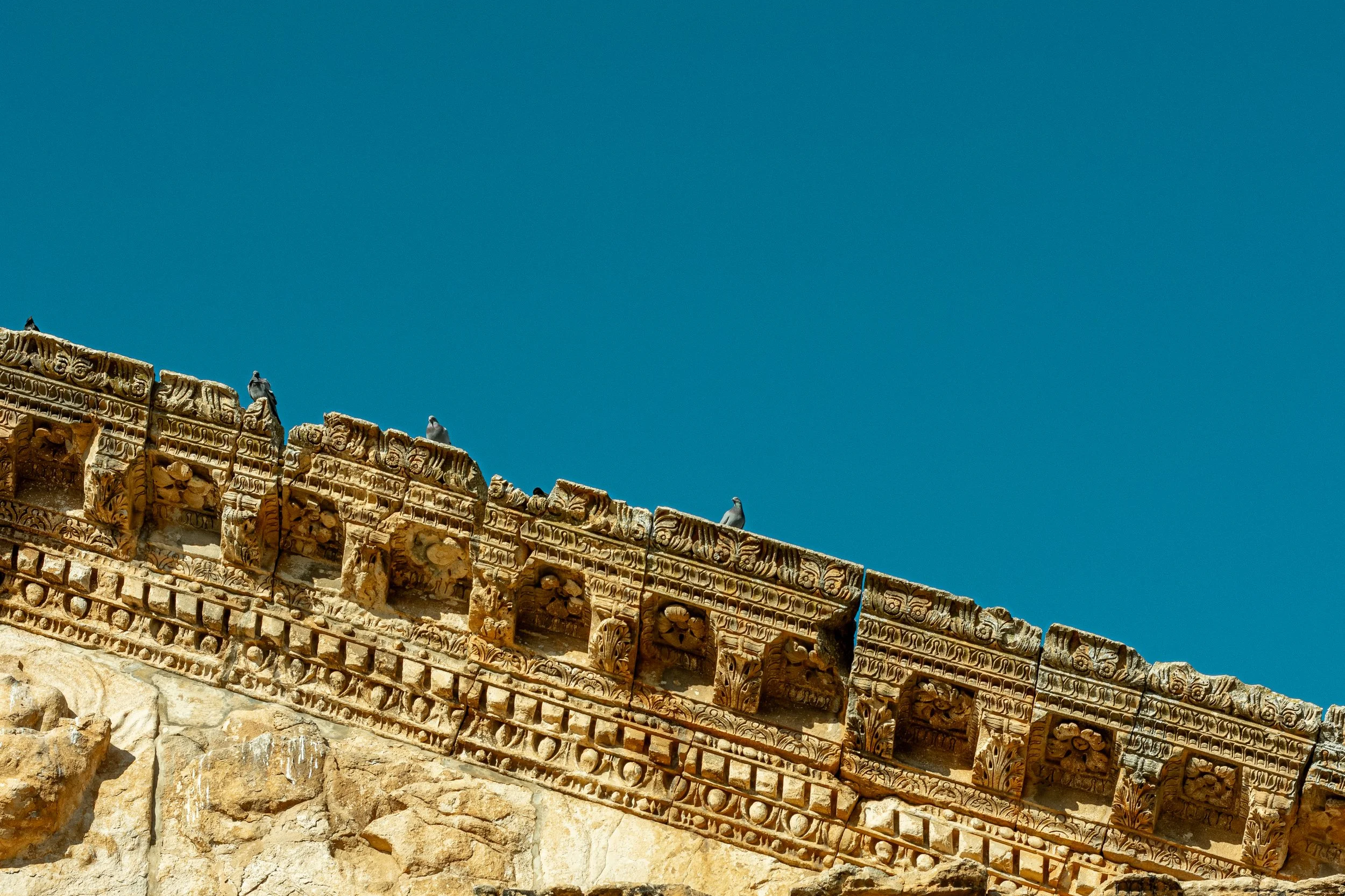 Ancient stone ruins with detailed carvings and sculptures, set against a clear blue sky, with three pigeons perched on top of the structure.