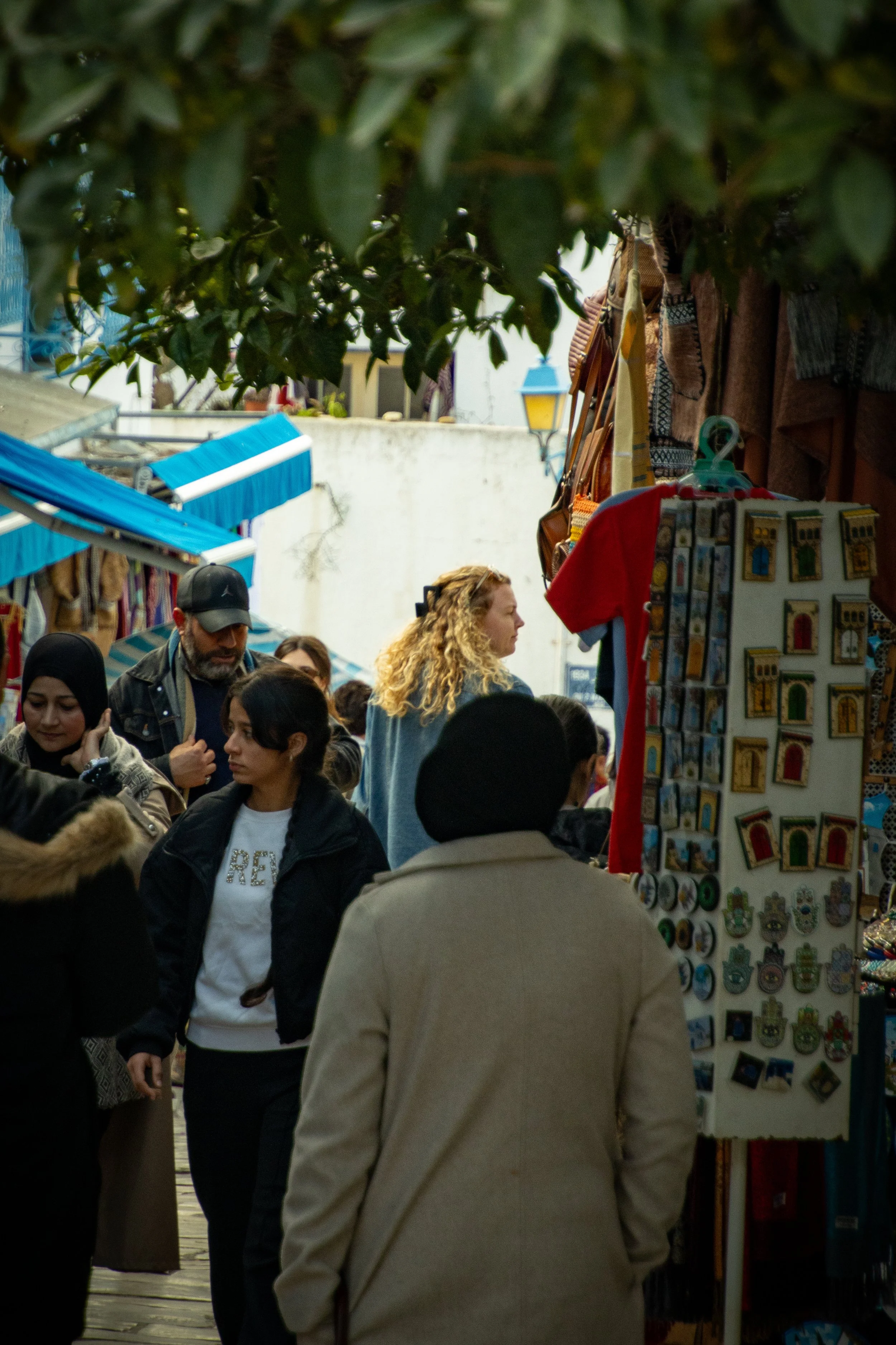 People shopping at an outdoor market with stalls selling jewelry and souvenirs. Some of the shoppers are looking at items, and a man wearing a black cap is visible. There are trees and colorful awnings in the background.
