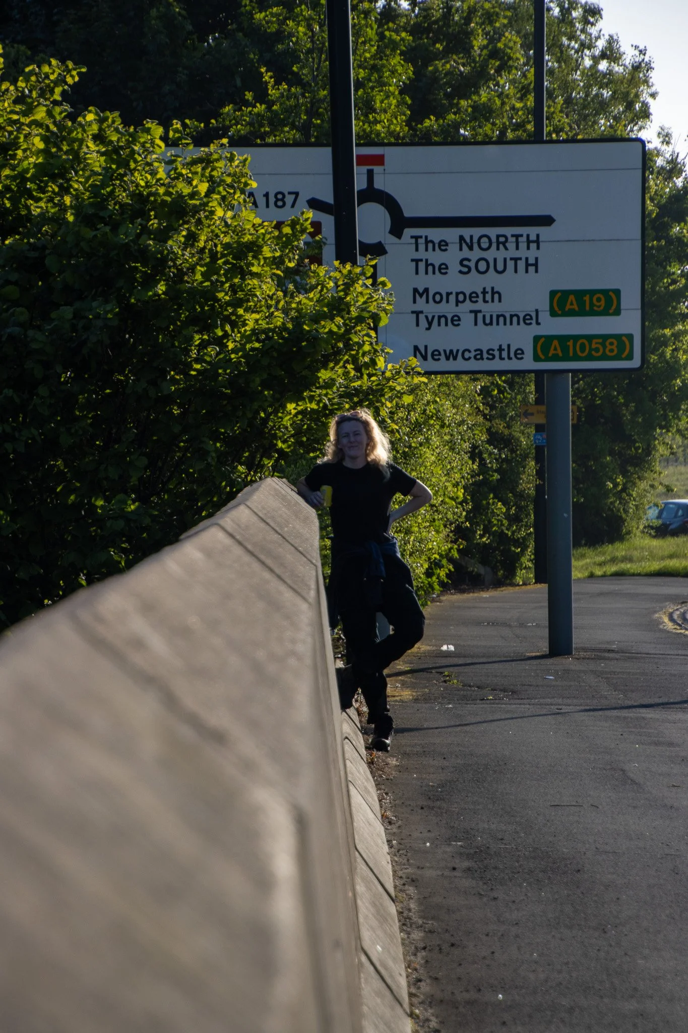A woman standing by a concrete barrier beside a street, with a street sign overhead indicating directions to The North, The South, Morpeth, Tyne Tunnel, and Newcastle. There are trees and a car in the background, and she is smiling with her hands on 