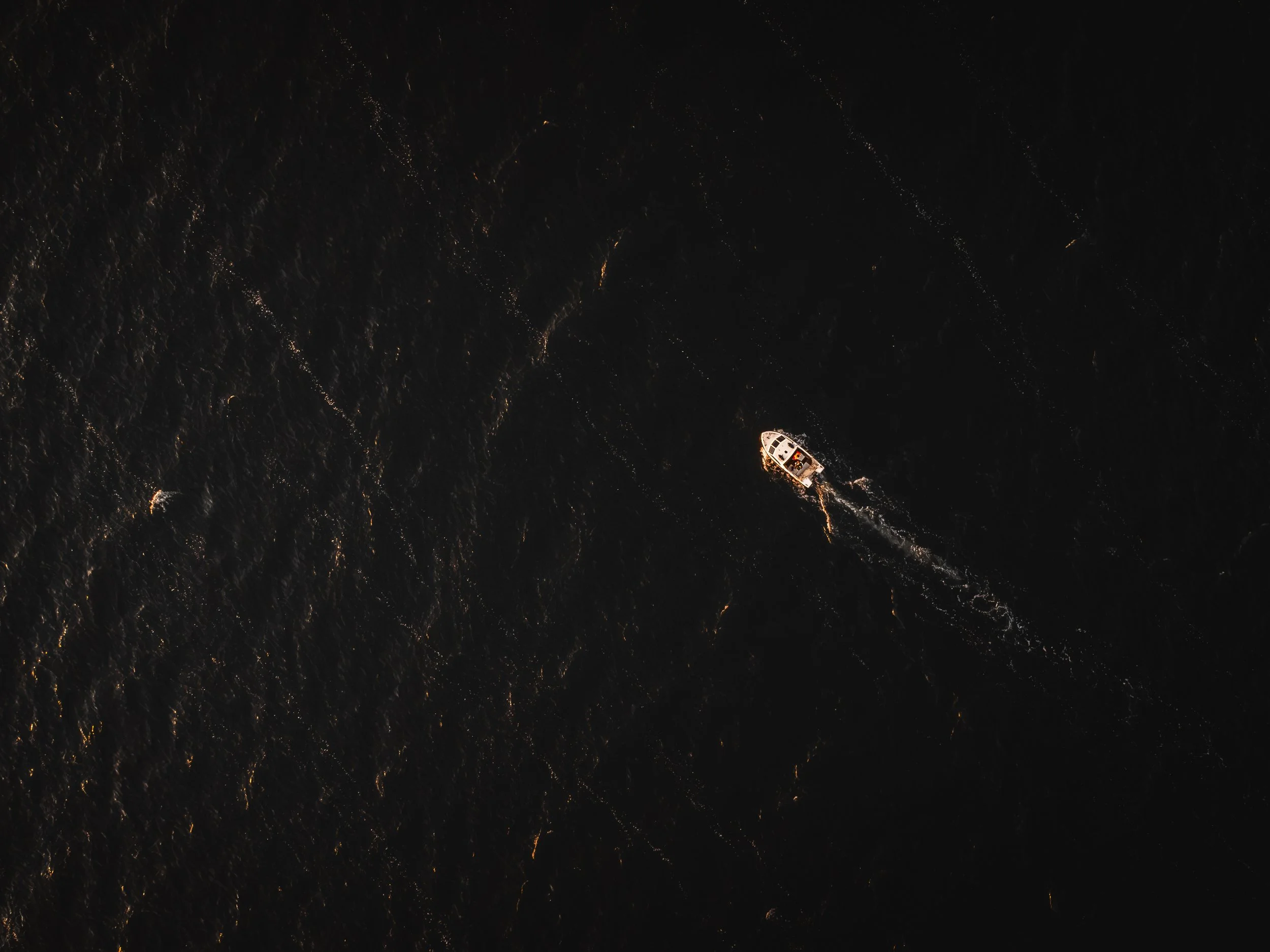 An aerial view of a dark ocean at night with a small boat floating on the water, illuminated by nearby lights.