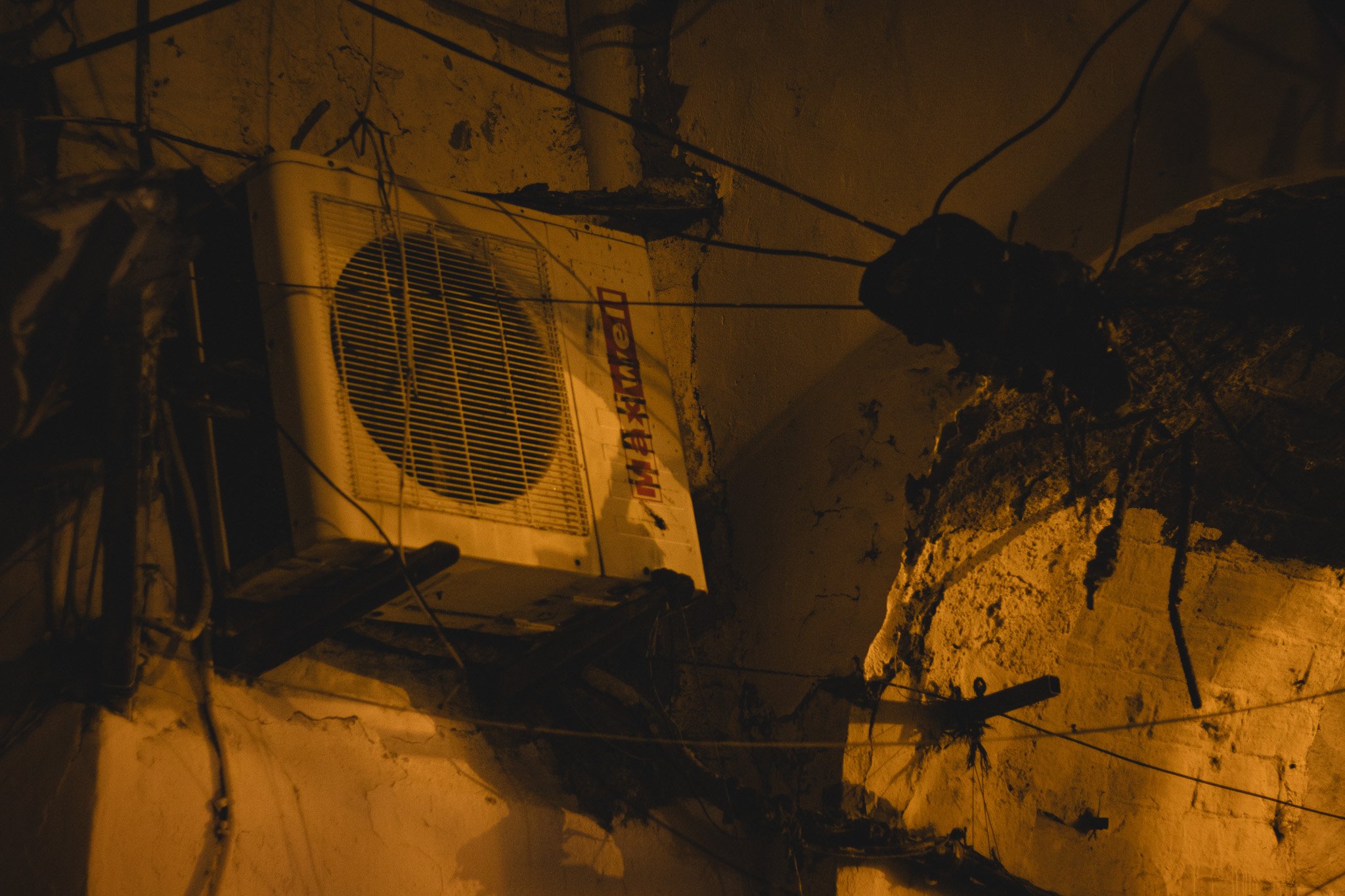 Nighttime street scene showing an air conditioning unit on a wall with electrical wires and a tree casting shadows.