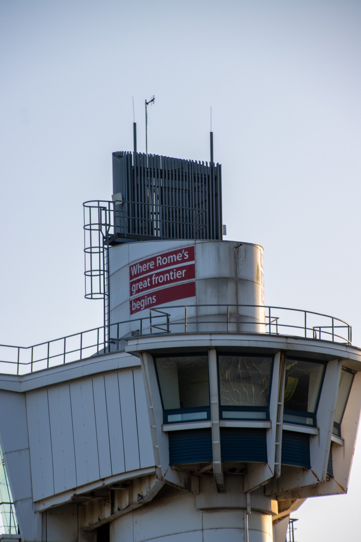 The control tower of an airport with a partially cloudy sky in the background, featuring antennas and a sign.