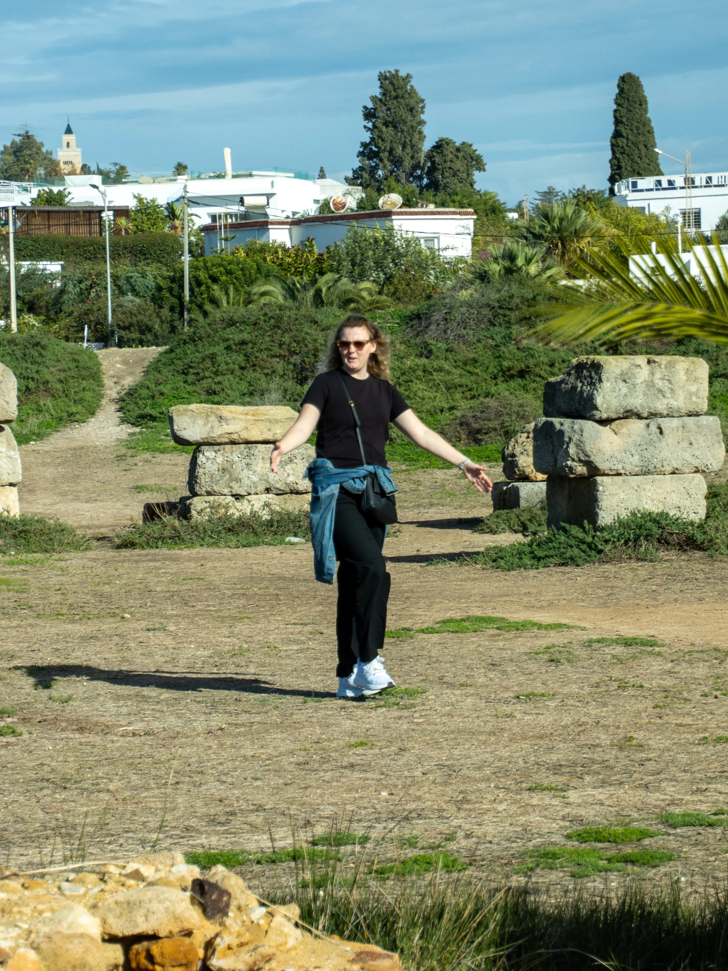 A woman with curly hair, wearing sunglasses, a black t-shirt, and black pants, walking outdoors on a dirt path surrounded by plants and rocks, with buildings and trees visible in the background under a cloudy sky.