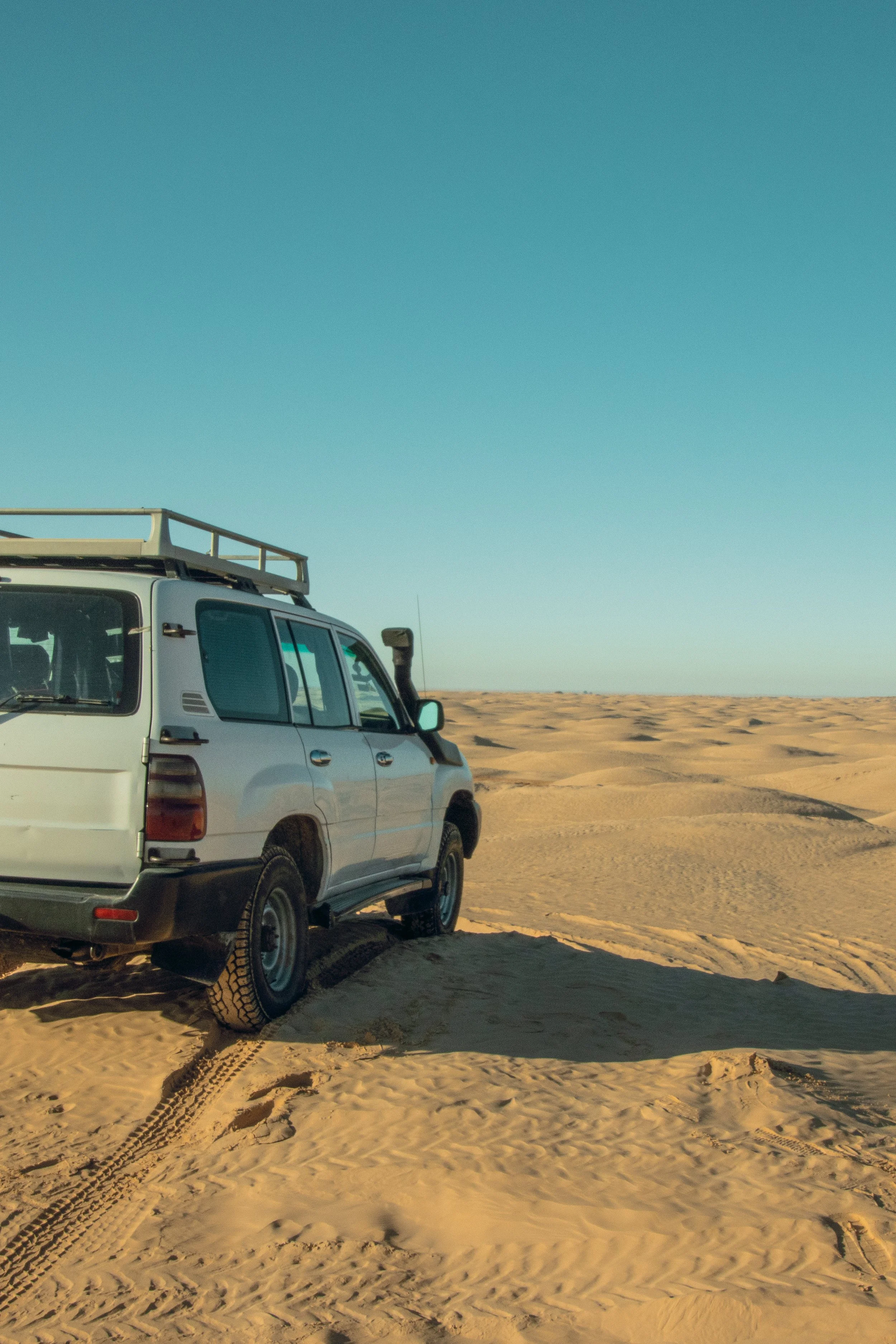 A white SUV vehicle parked on a sandy desert landscape under a clear blue sky.