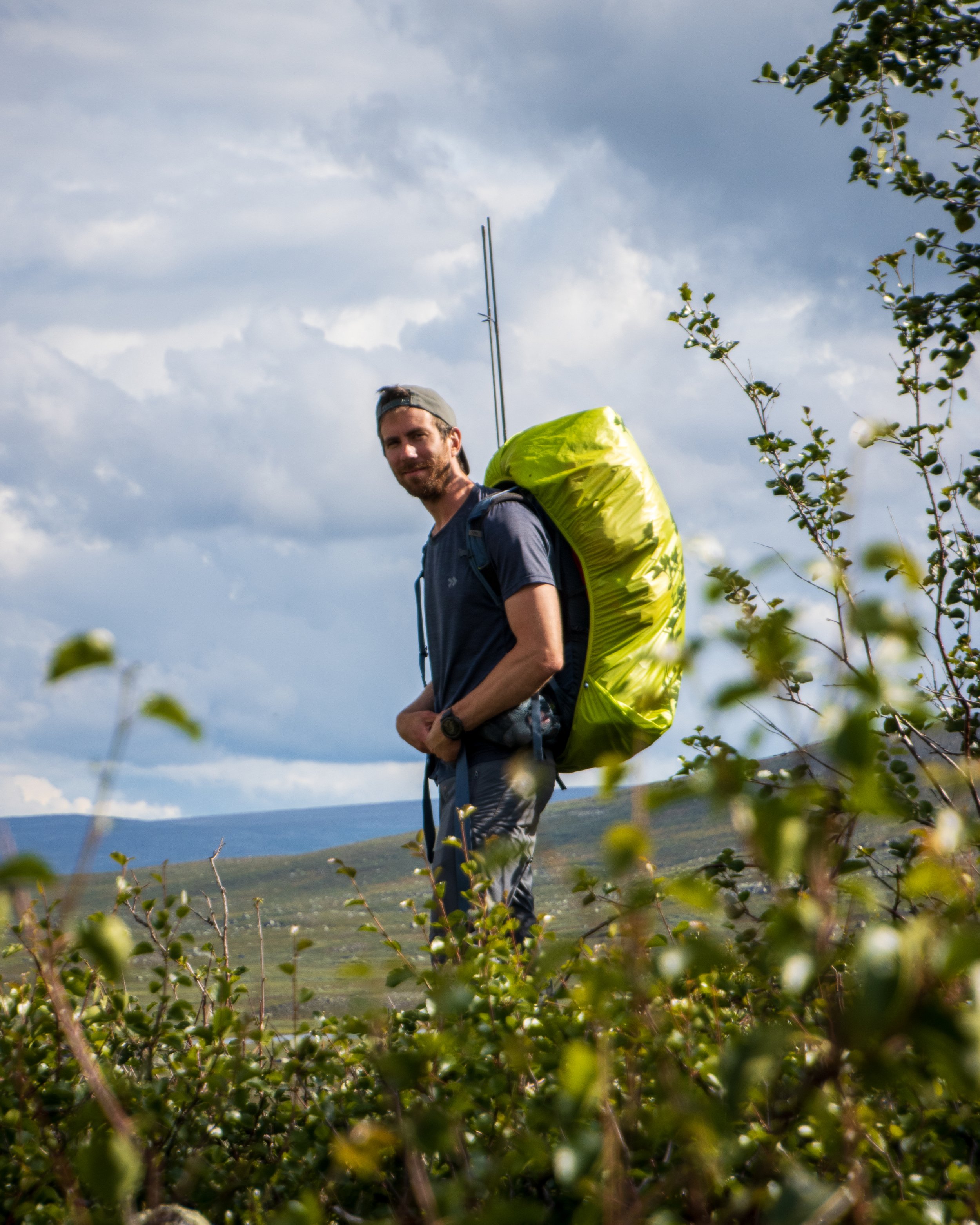 A man with a beard, wearing a gray cap backward, a dark shirt, and a watch, is hiking outdoors with a large yellow backpack. He is standing amidst dense bushes, with a scenic background of rolling hills, trees, and a cloudy sky.