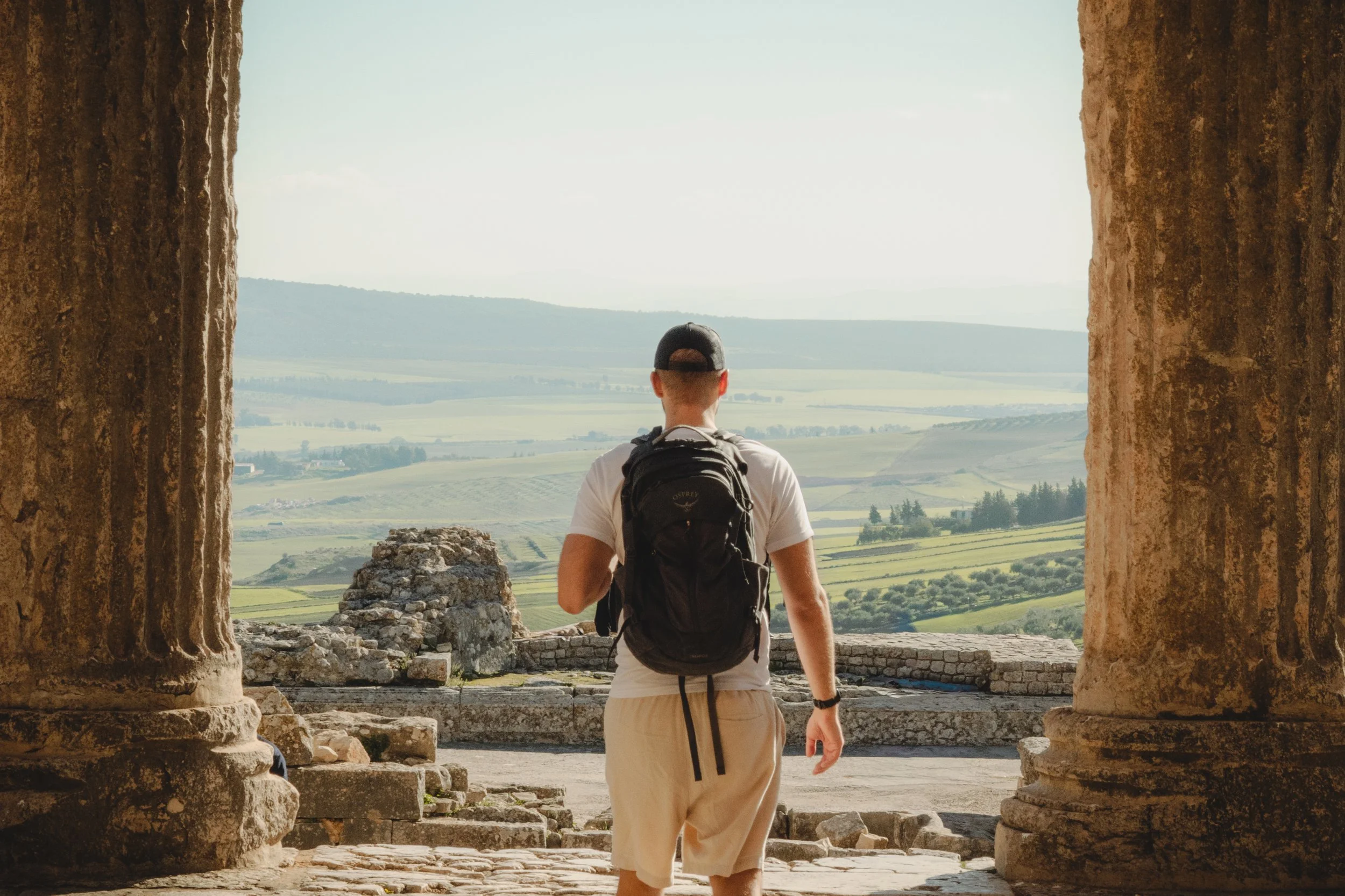 A man with a backpack walking through ancient stone ruins, viewing a scenic landscape of fields and hills in the distance, framed by large stone pillars.