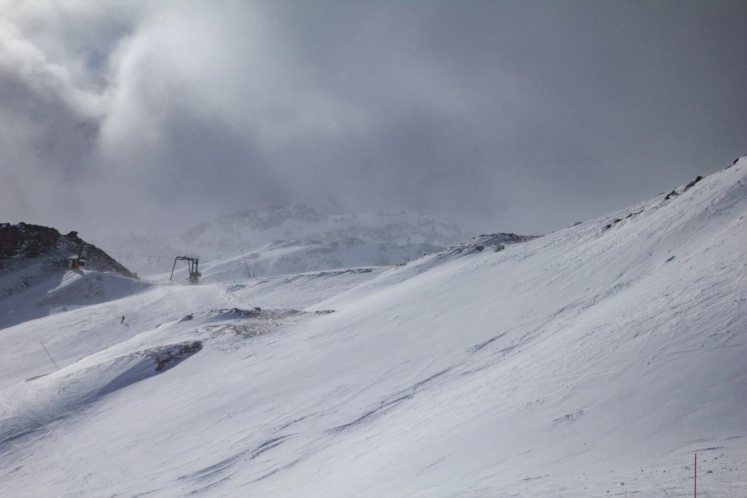 Snow-covered mountain slope with ski lift structures and a cloudy sky.