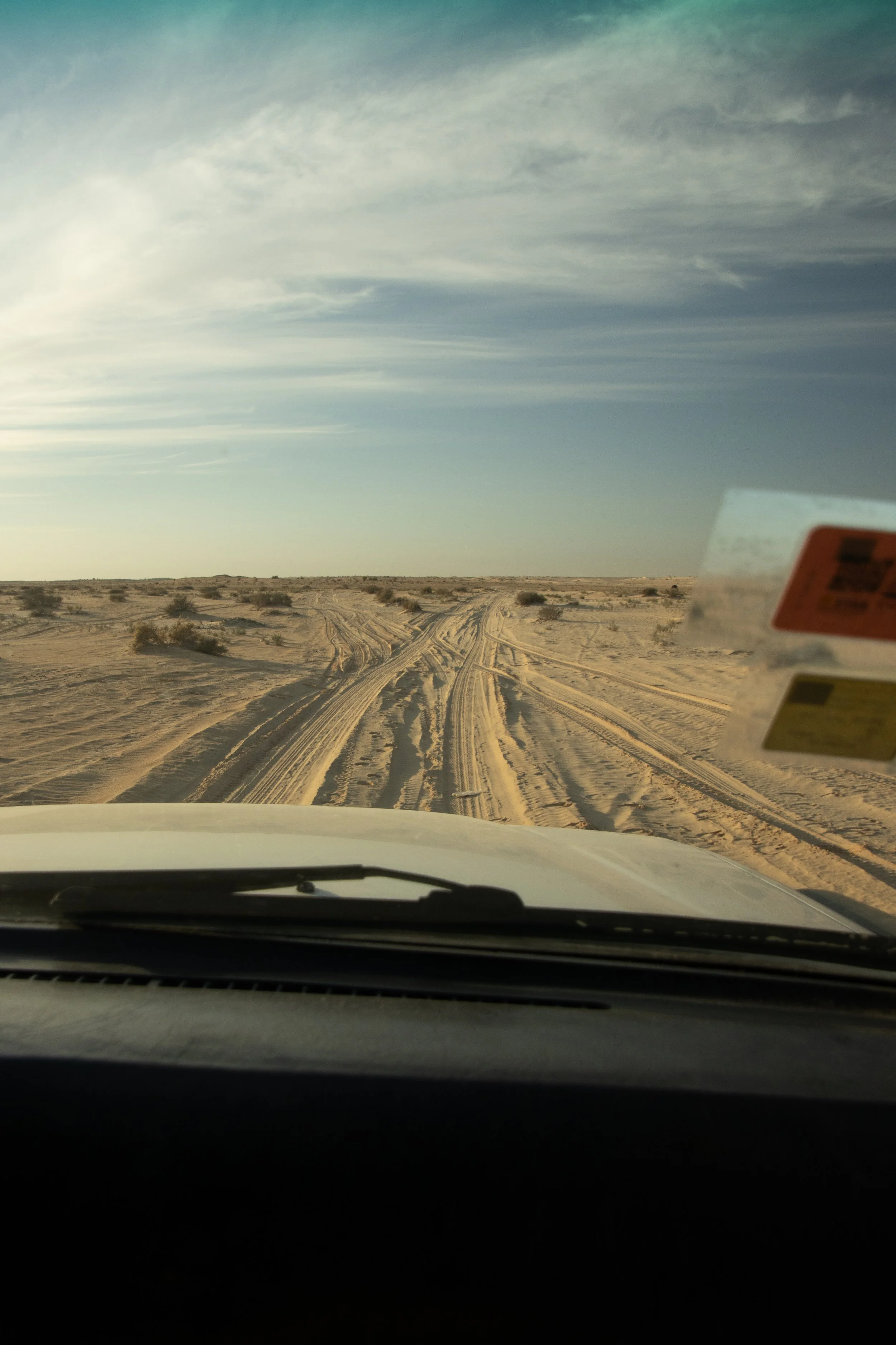 View through a car windshield of sandy desert terrain with tire tracks and small bushes, under a cloudy sky.
