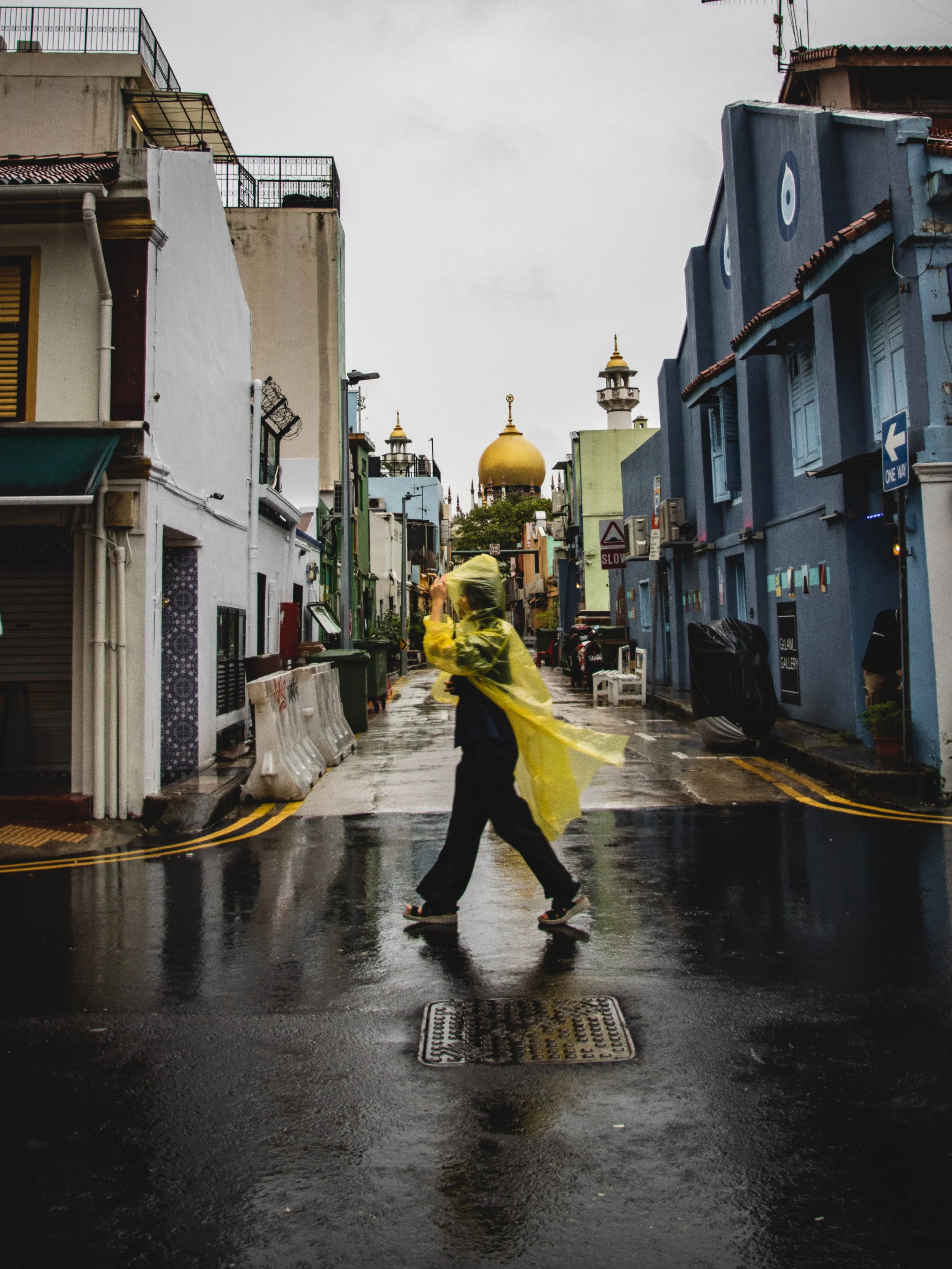 A person in a yellow raincoat is crossing a wet street while holding an umbrella in a city. The background features colorful buildings and a mosque with a gold dome and minarets, under an overcast sky.