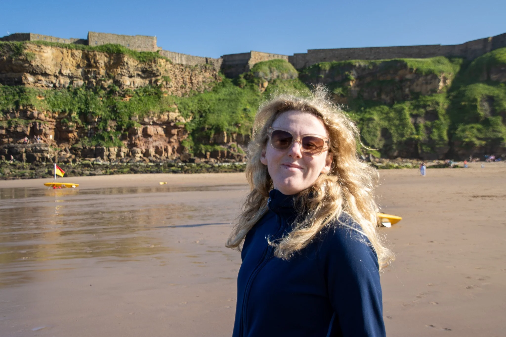A woman with curly blonde hair wearing sunglasses and a dark jacket stands on a sandy beach with a castle wall and green hillside in the background during daytime.