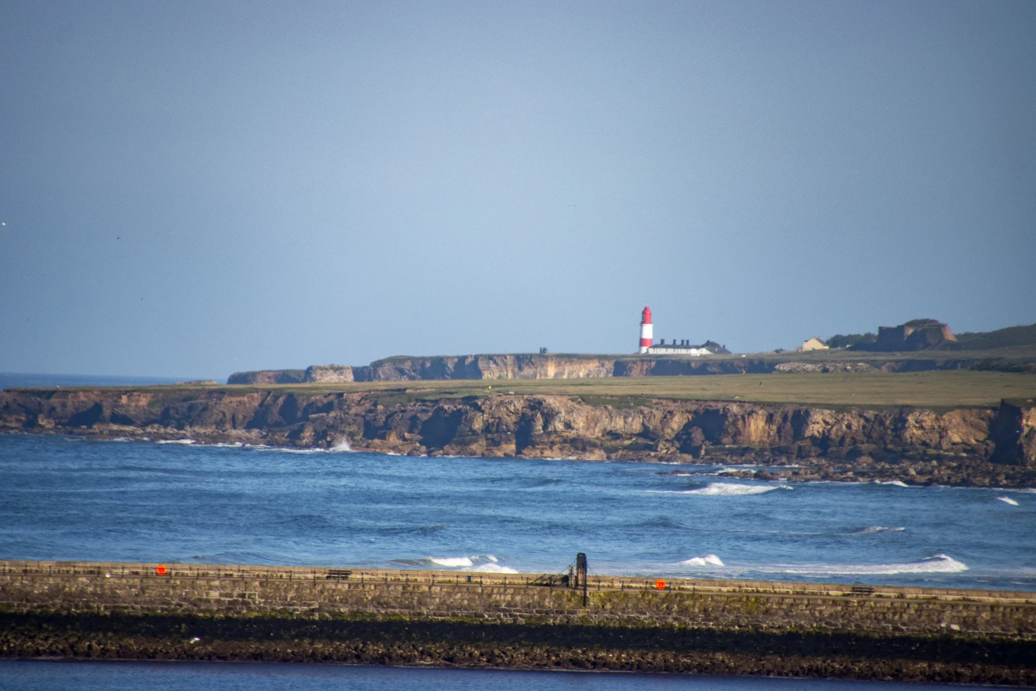 Scenic view of rocky coastline with cliffs, ocean waves, and a lighthouse on a hill in the distance under a clear sky.