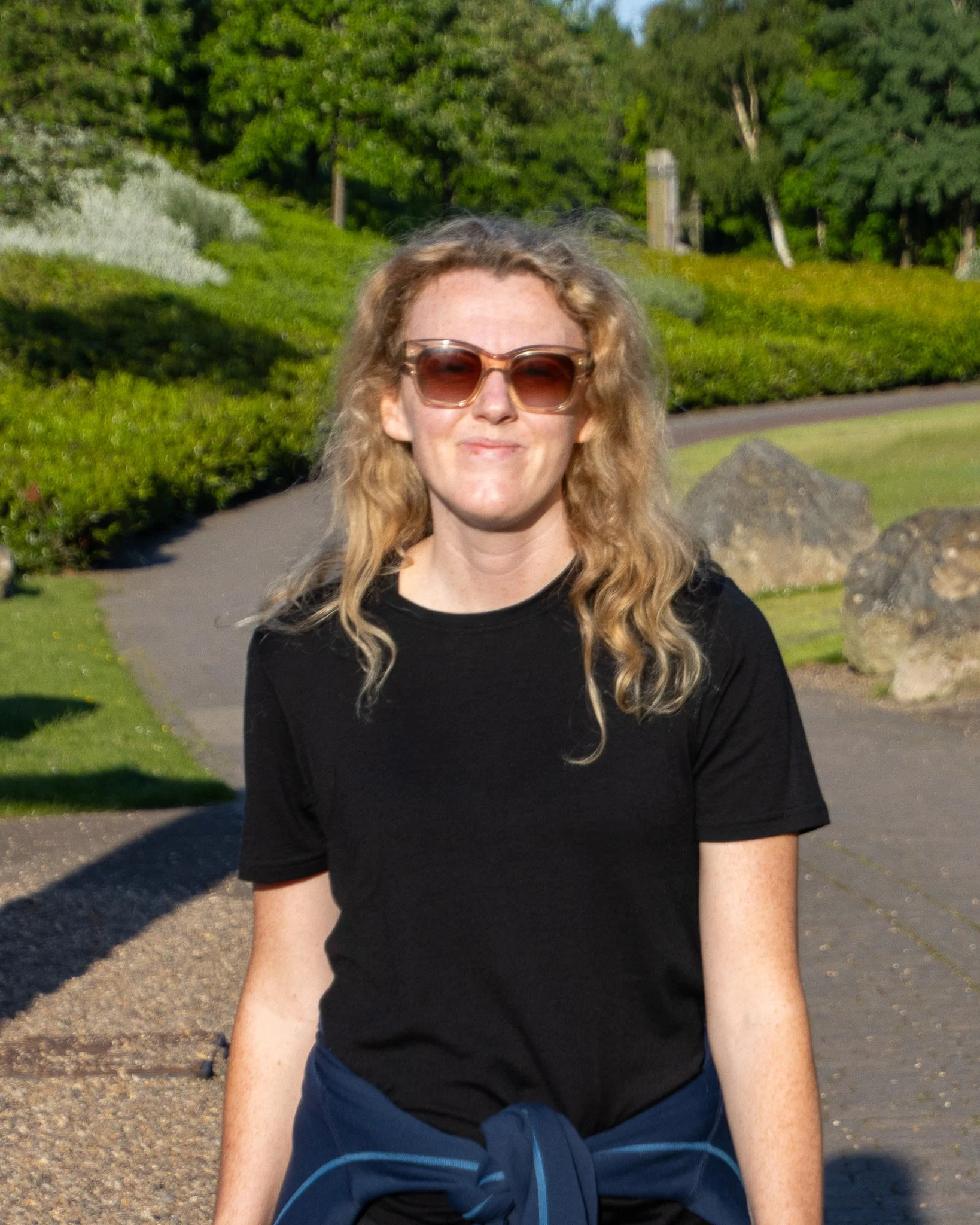 A woman with curly blonde hair wearing sunglasses and a black shirt, sitting outdoors on a sunny day, with a grassy park and trees in the background.