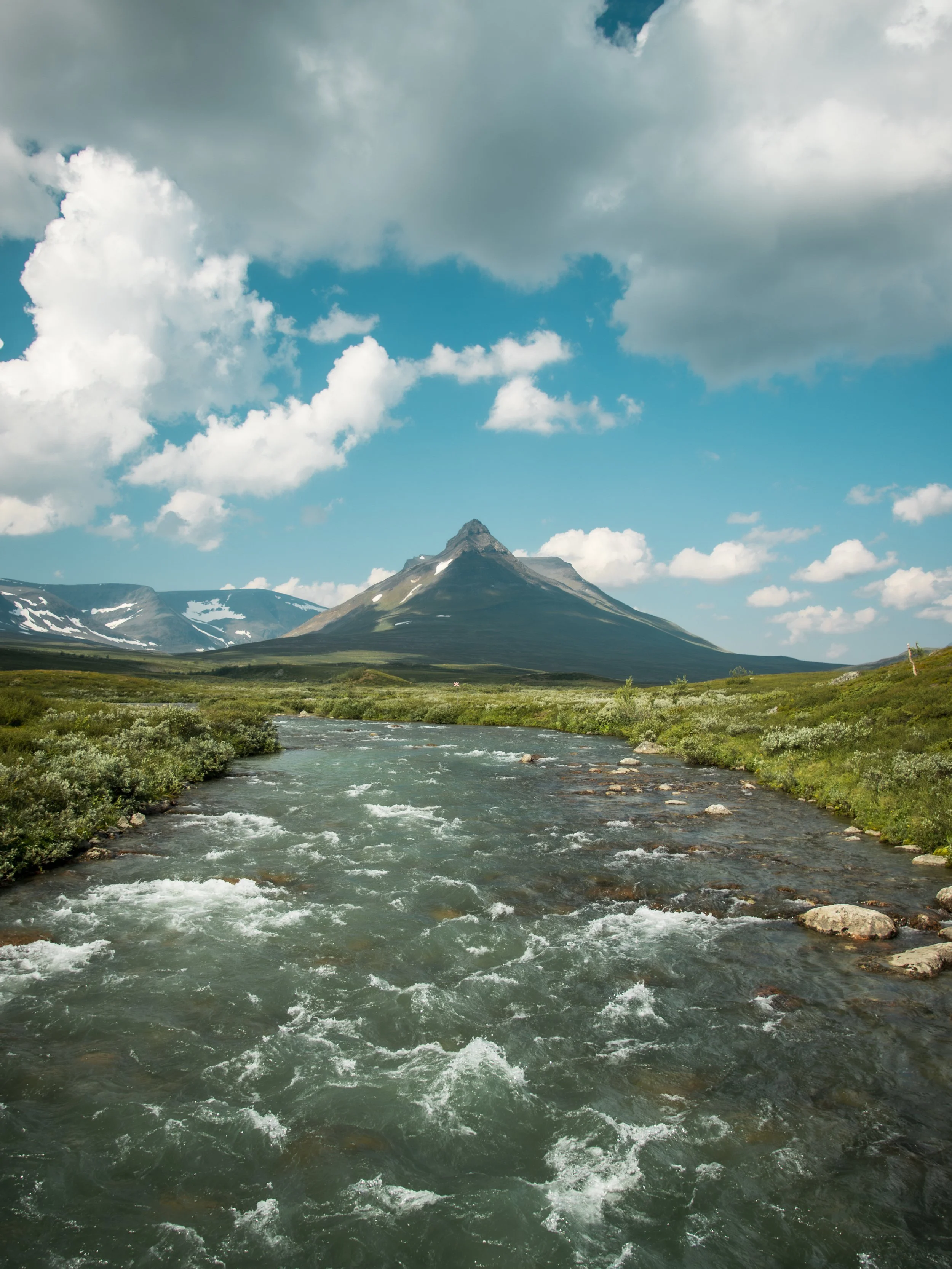 A river flowing through a green landscape with a mountain in the background and a partly cloudy sky.
