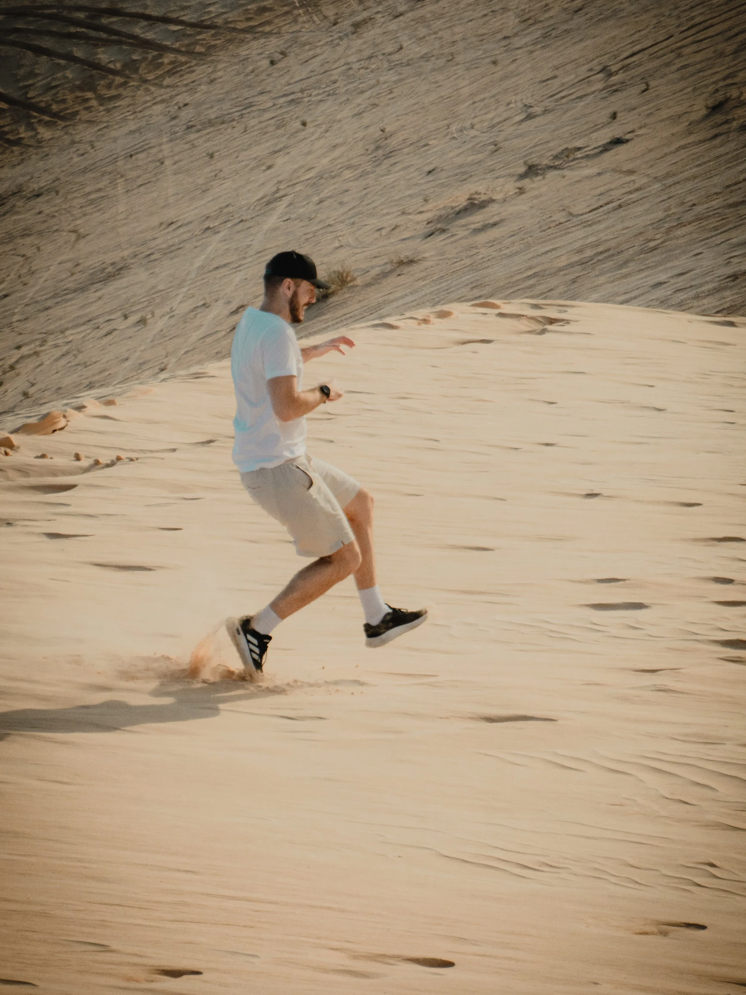 A man running in a desert with sand dunes, wearing a white t-shirt, shorts, sneakers, and a cap.