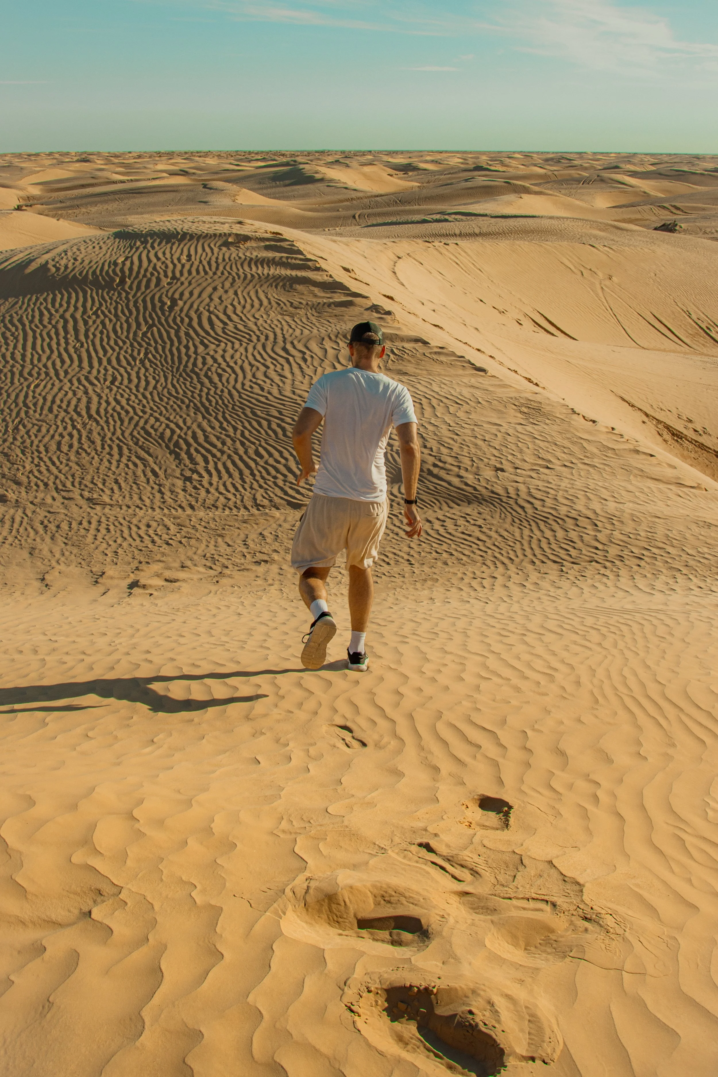 A man running in a desert with sand dunes, footprints, and a clear sky.