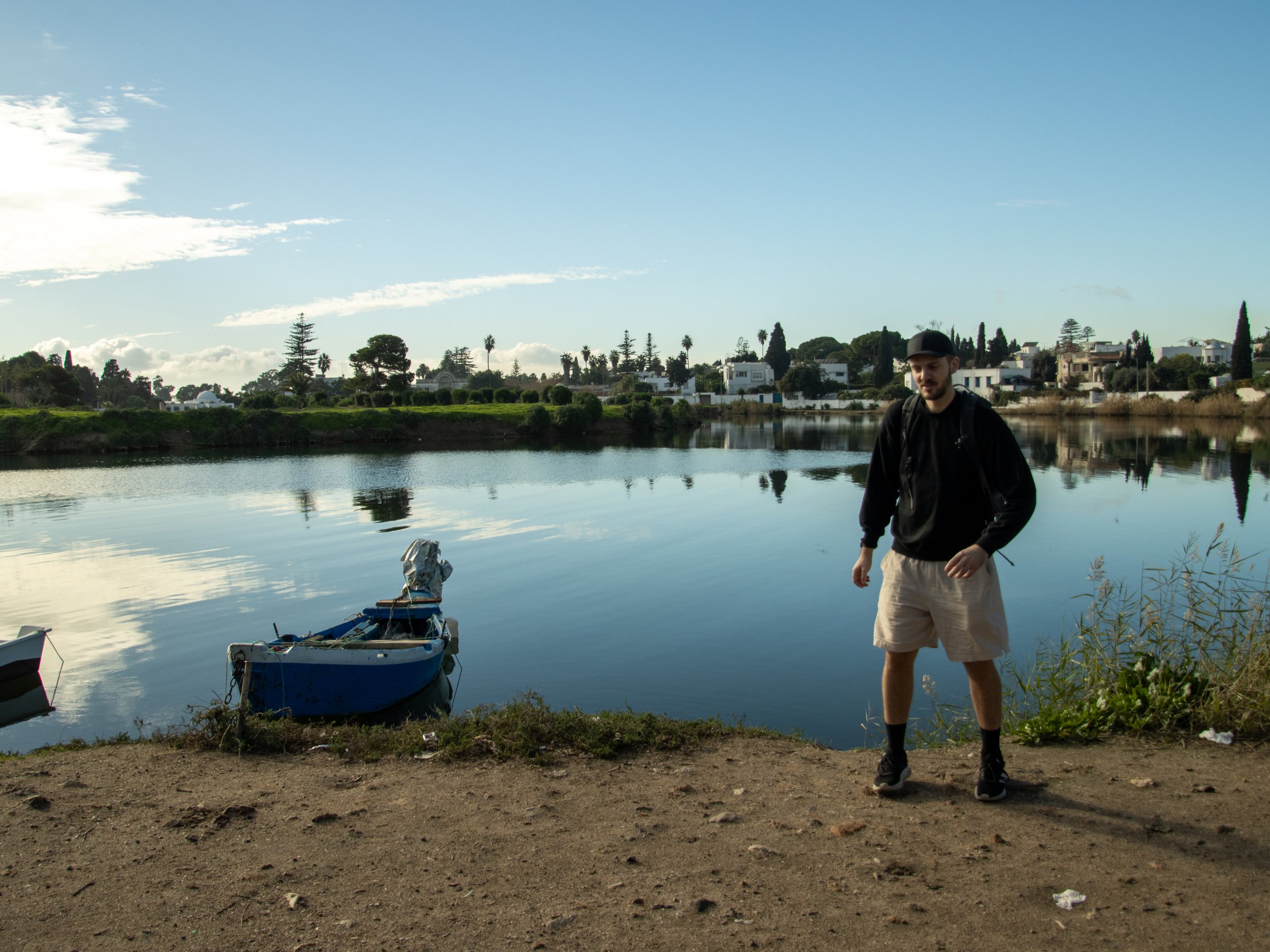 A man standing near the edge of a lake with small boats floating nearby, houses and trees across the water, reflecting in the lake, under a partly cloudy sky.