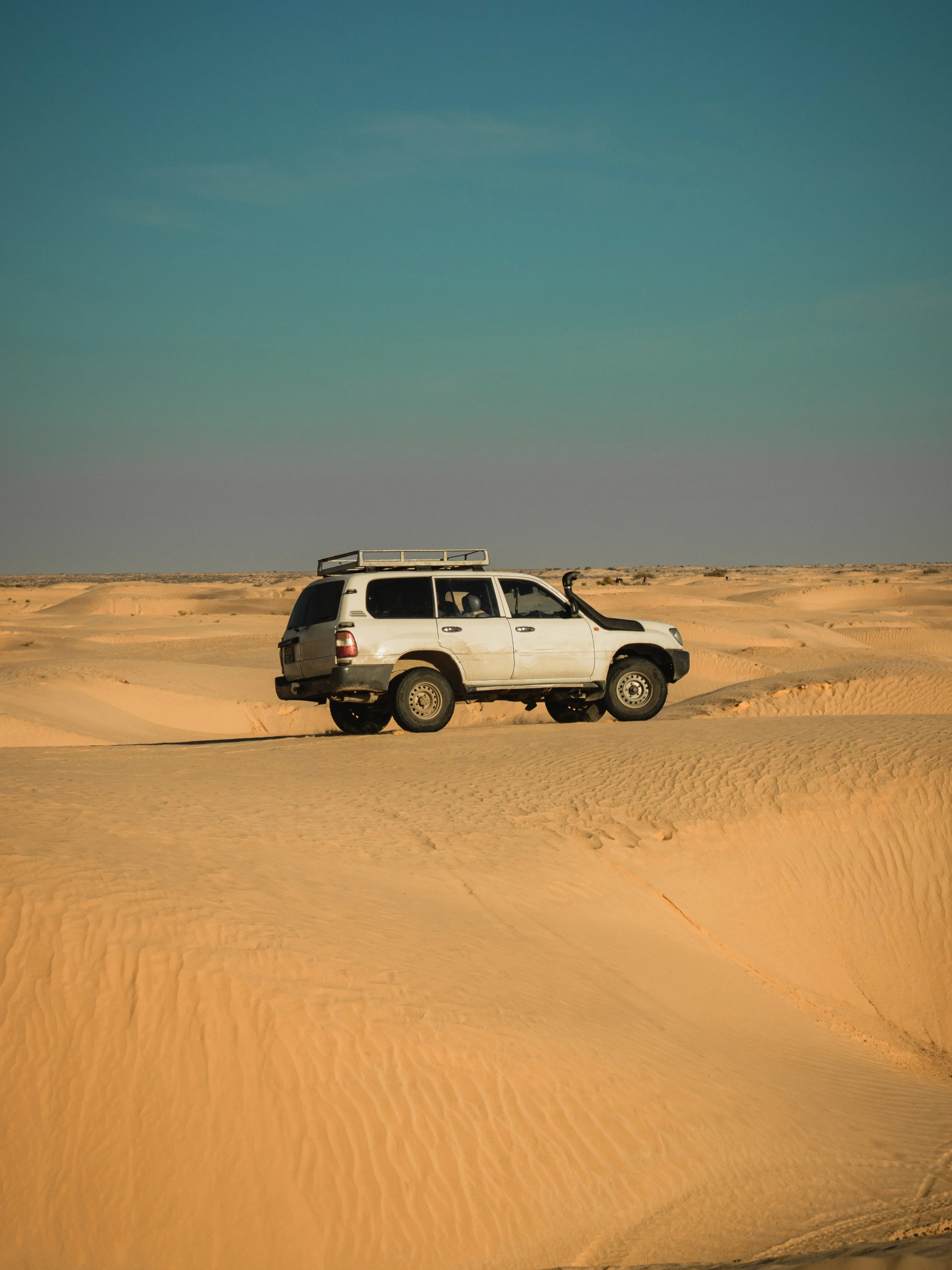 A white SUV vehicle parked on sandy desert dunes with a clear blue sky in the background.