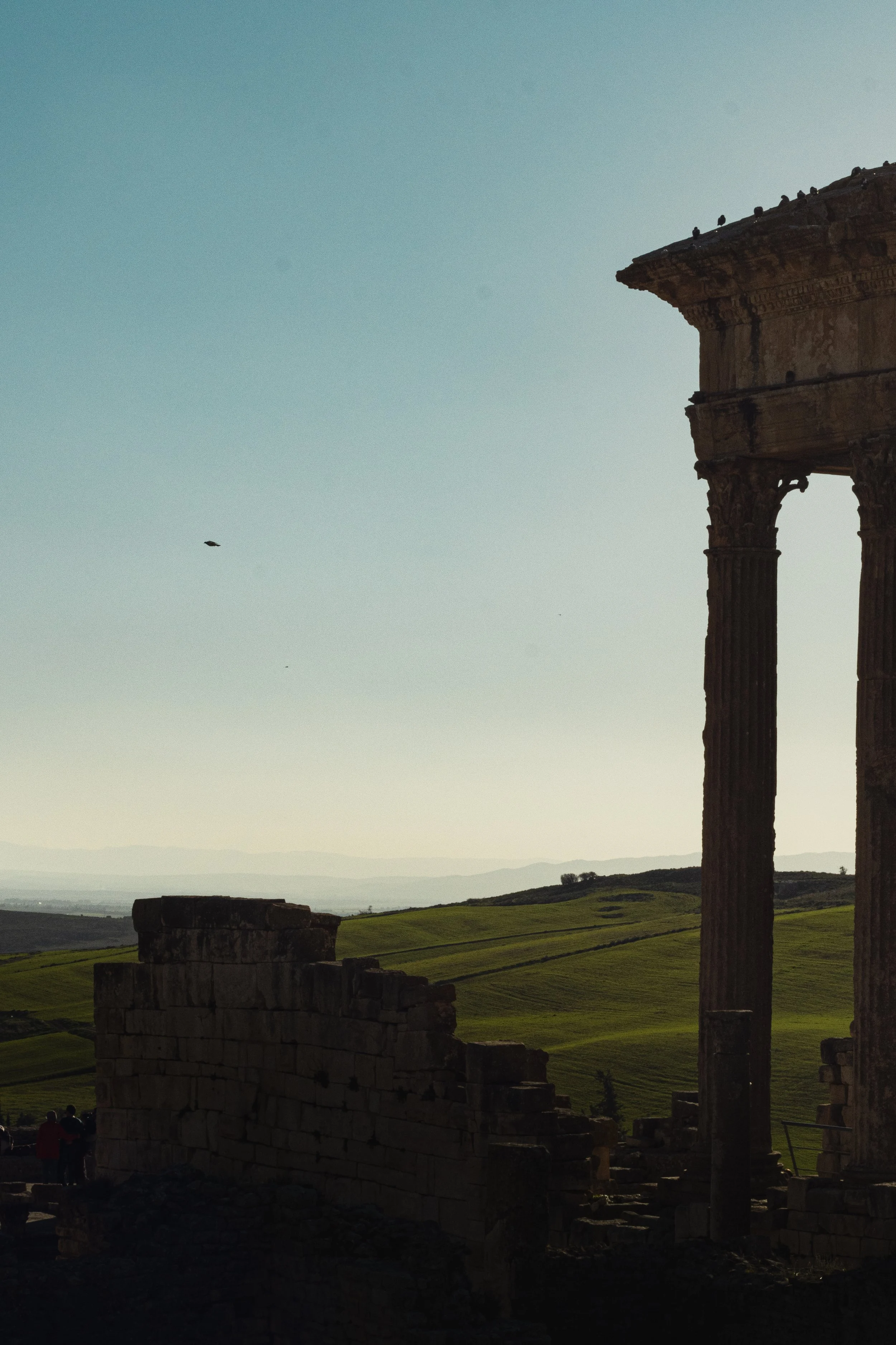 Ancient ruins of stone columns and walls with a scenic landscape of green rolling hills in the background, under a clear sky.