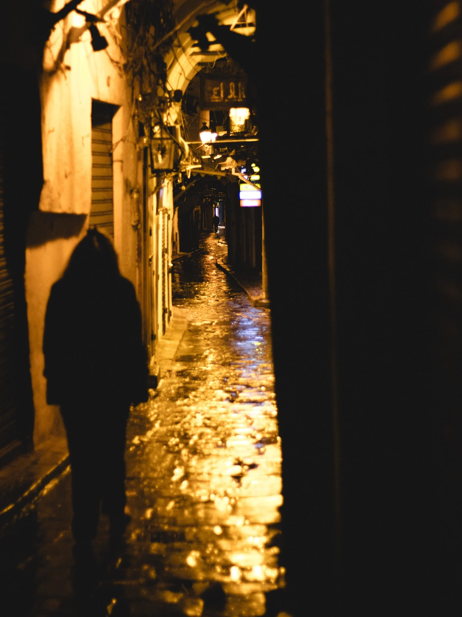 Night scene of a narrow alleyway reflecting streetlights on wet cobblestone pavement; a silhouette of a person walking away is visible in the distance.